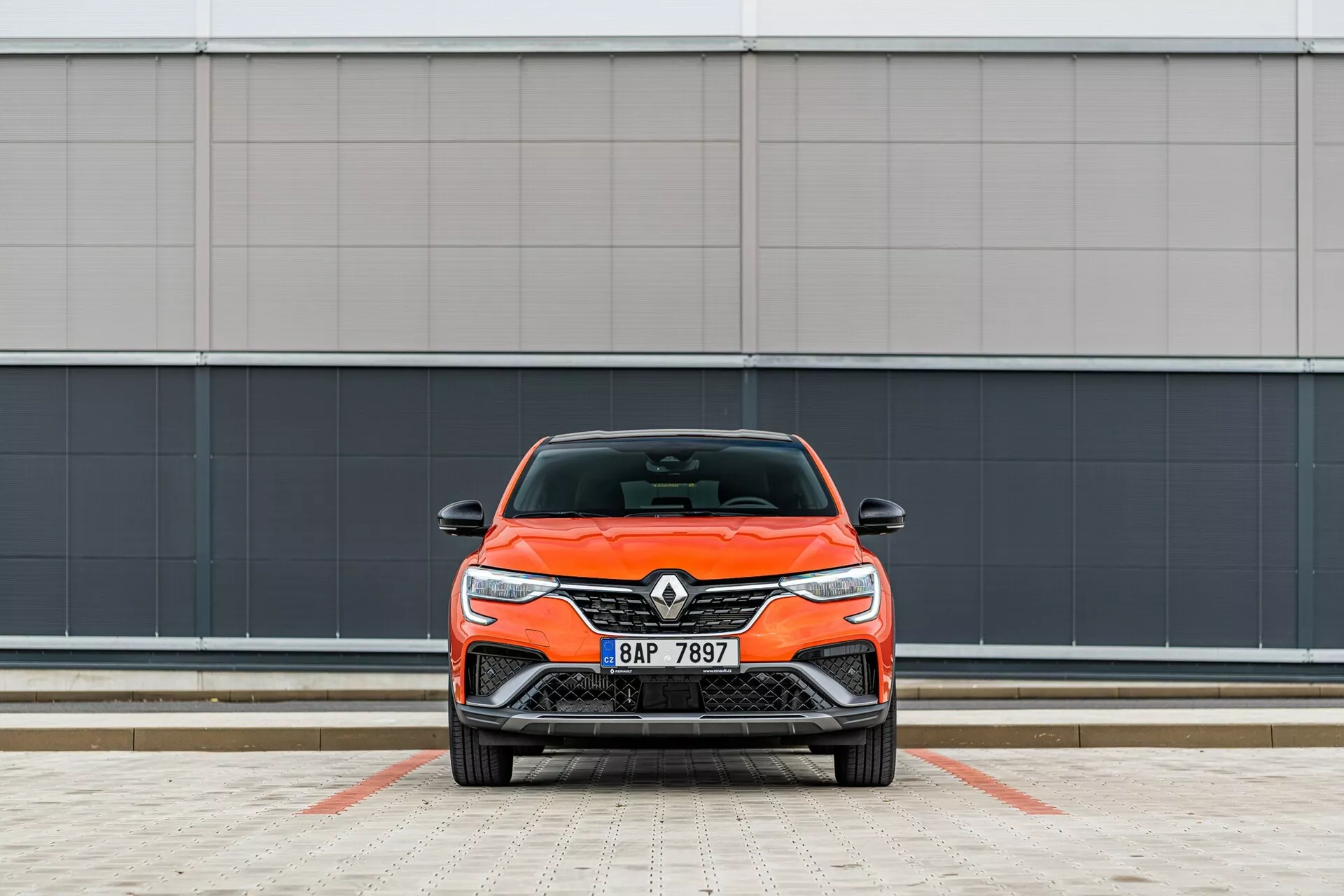 Čelný pohľad na jasno oranžové auto Renault, zvýrazňujúce jeho moderný dizajn s elegantnými svetlometmi a prednou mriežkou, zaparkované kolmo ku kamere pred minimalistickým sivým pozadím na vyznačenom parkovisku.