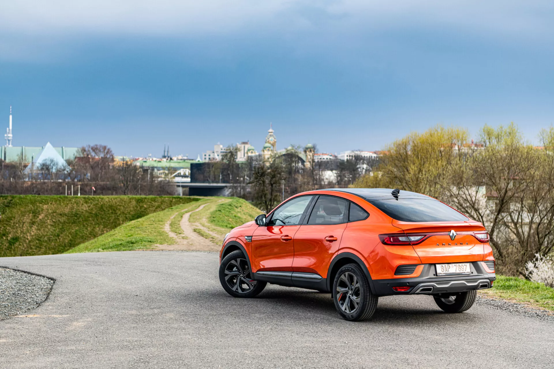 Oranžové kompaktné crossover SUV Renault Arkana 2021 pod pravým uhlom ukazujúce profil a čiastočný zadný výhľad, umiestnené na prírodnom pozadí s mäkkým osvetlením zdôrazňujúcim jeho športový kupéový elegantný dizajn a moderné vlastnosti.