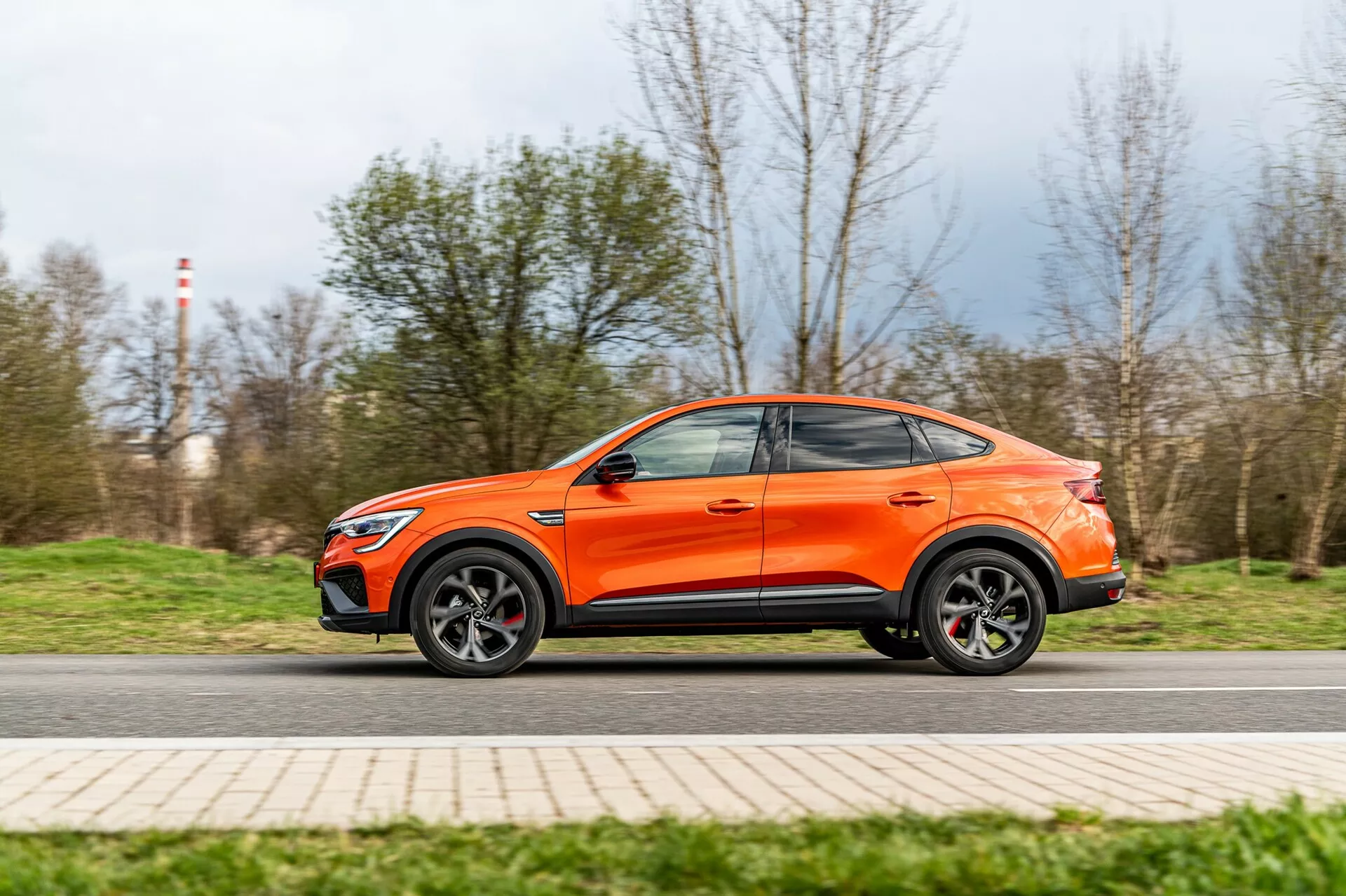 Dynamický bočný pohľad na oranžový crossover SUV Renault Arkana 2021 v pohybe, zachytený z nízkeho uhla zdôrazňujúceho svetlú výšku a športový postoj. Viditeľná je celá dĺžka vozidla, ukazujúca dizajnové línie, zliatinové kolesá, kontúrované bočné dvere a strechu vo štýle kupé. Mierny pohybový rozmaz na kolesách a pozadí naznačuje miernu rýchlosť, pričom auto je ostré. Obrázok zdôrazňuje moderný a aerodynamický dizajn vozidla.