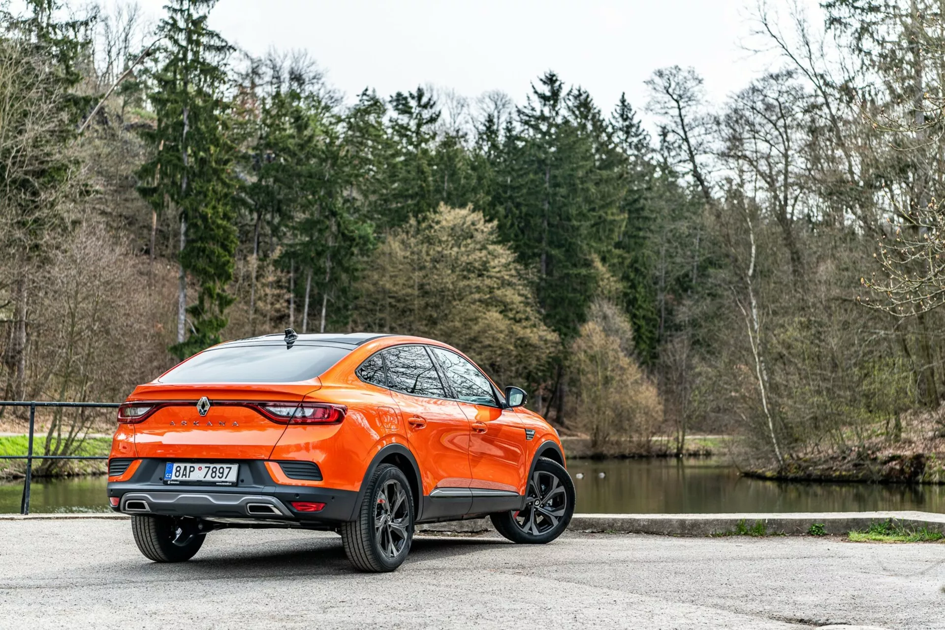 Renault Arkana 2021 zobrazený zo zadného trochštvrťového uhla, podrobný pohľad na bočný a zadný profil, predstavujúci moderný crossover/SUV so zvýšenou svetlou výškou a športovou líniou strechy typu kupé. Výrazná oranžová karoséria vyniká na pozadí stromov a jazera, zdôrazňujúc viacramenné dizajnové liatinové kolesá, zadné LED svetlá, zadný spojler, anténu typu žraločej plutvy, centrálne umiestnený emblém, dvojité výfukové koncovky, lesklý povrch laku a zadné zatmavené okná. Rozptýlené prírodné osvetlenie zlepšuje viditeľnosť dobre udržiavaného exteriéru, čo naznačuje príťažlivosť pre spotrebiteľov s aktívnym životným štýlom, zaparkované na dláždenom povrchu.