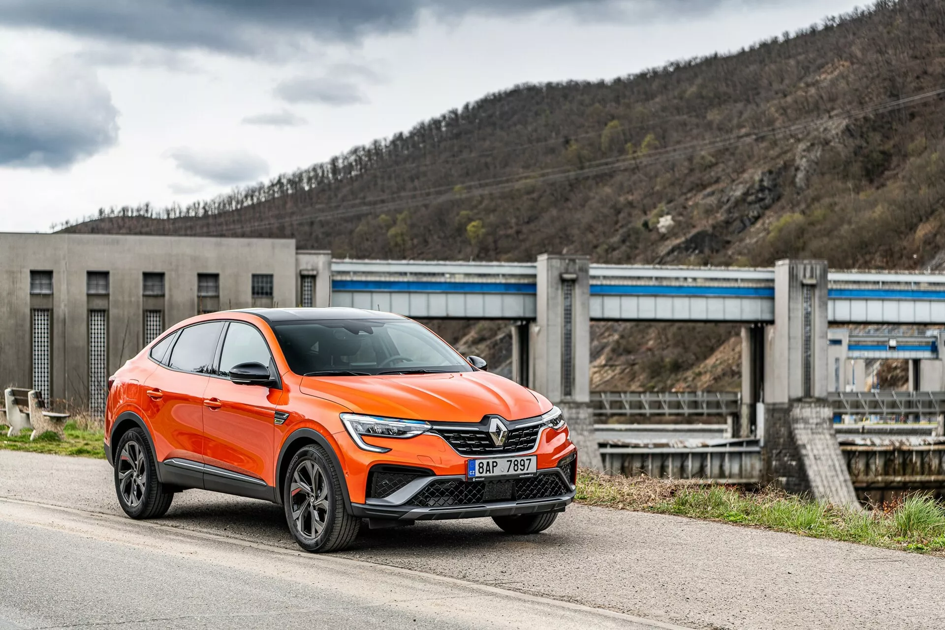 Moderný jasne oranžový crossover SUV Renault Arkana 2021 je zaparkovaný pri ceste, fotografovaný z predno-bočného uhla zameraného na stranu vodiča. Obrázok ukazuje prednú mriežku vozidla s veľkým emblémom, elegantné uhlové LED svetlomety, denné svetlá, prívody vzduchu a pravdepodobne senzory asistenčných systémov vodiča na spodnom nárazníku, výrazné blatníky s viacramennými zliatinovými kolesami a zrkadlá s integrovanými smerovými svetlami. Vozidlo, klasifikované ako SUV, má zvýšenú polohu s väčšou svetlou výškou a jemne klesajúcu strechu v štýle kupé. V pozadí je viditeľná infraštruktúra pripomínajúca priehradu alebo záplavový systém, ktorá umiestňuje vozidlo do priemyselného alebo vidieckeho prostredia. Záber je urobený z mierne nízkeho uhla, čo zdôrazňuje výšku a postoj vozidla.