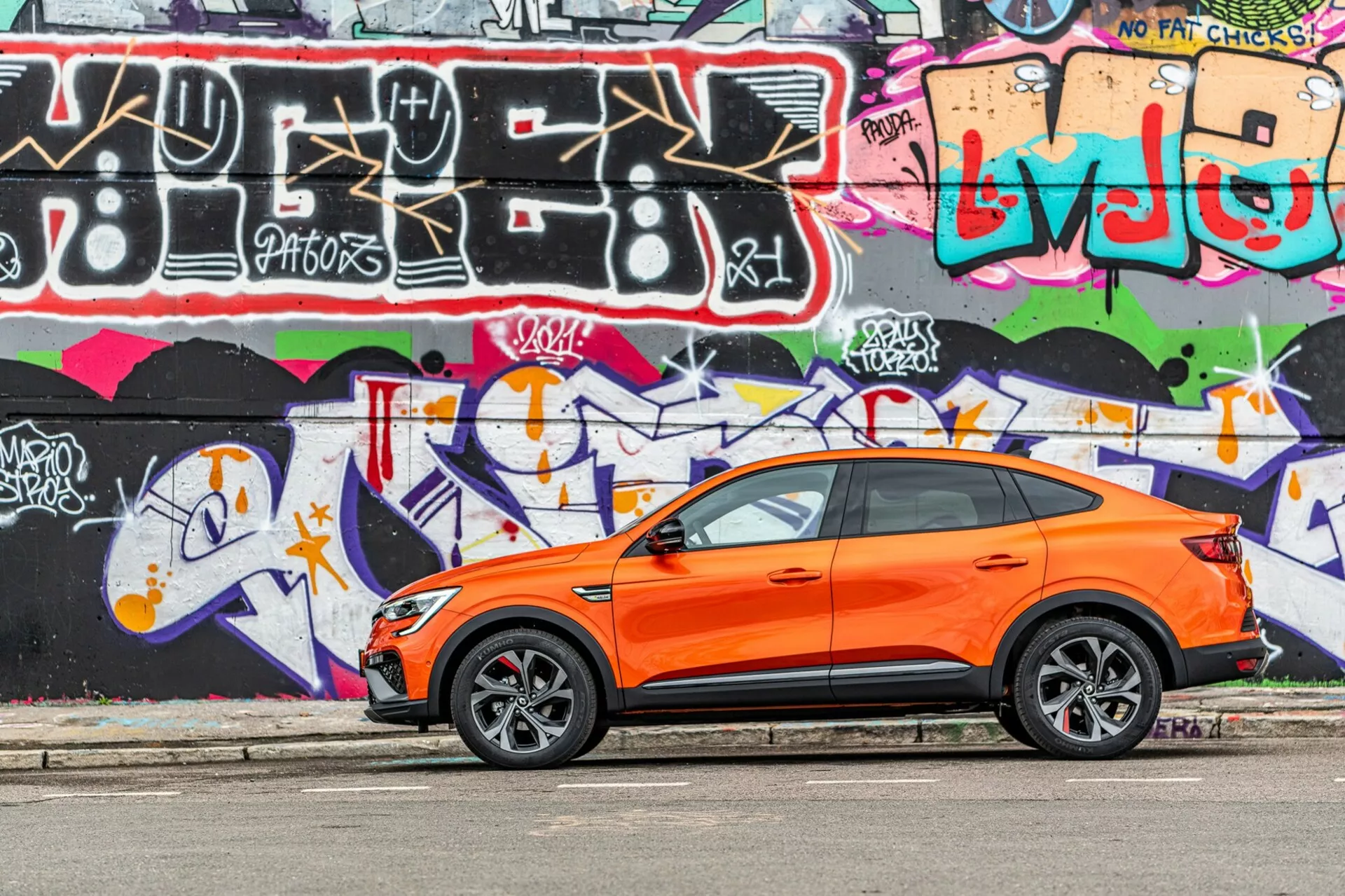 Obrázok zobrazuje žiarivo oranžový crossover SUV Renault Arkana 2021 zaparkovaný paralelne s ulicou s pestrofarebnou graffiti stenou v pozadí. Fotografia zachytená z bočného uhlu zvýrazňuje športový a moderný dizajn vozidla s viditeľnými prednými a zadnými dverami vodiča, bočnými oknami, bočnými zrkadlami a klesajúcou líniou strechy. Štýlové zliatinové kolesá s nízkoprofilovými pneumatikami naznačujú zameranie na výkonnosť na ceste. Kľučky na predných dverách naznačujú systém bezkľúčového vstupu a štartu. Obrázok bol nasnímaný za denného svetla, zdôrazňujúc kontrast oranžovej karosérie oproti graffiti a ukazujúc dobre udržiavanú, ostrú karosériu vozidla v mladistvom, mestskom prostredí.