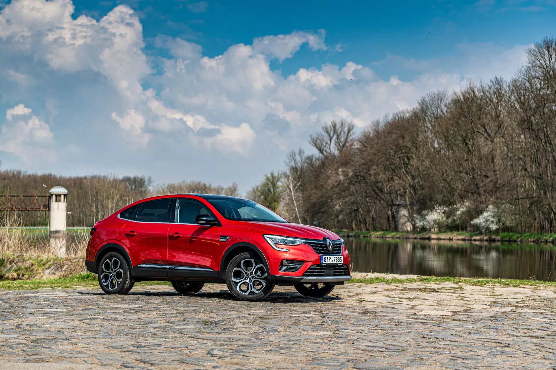 Červené SUV Renault Arkana 2021 zaparkované na kamennej dlažbe s vodou a stromami na pozadí pod čiastočne zamračenou oblohou, bočný profil zdôrazňuje elegantný dizajn a dynamické funkcie