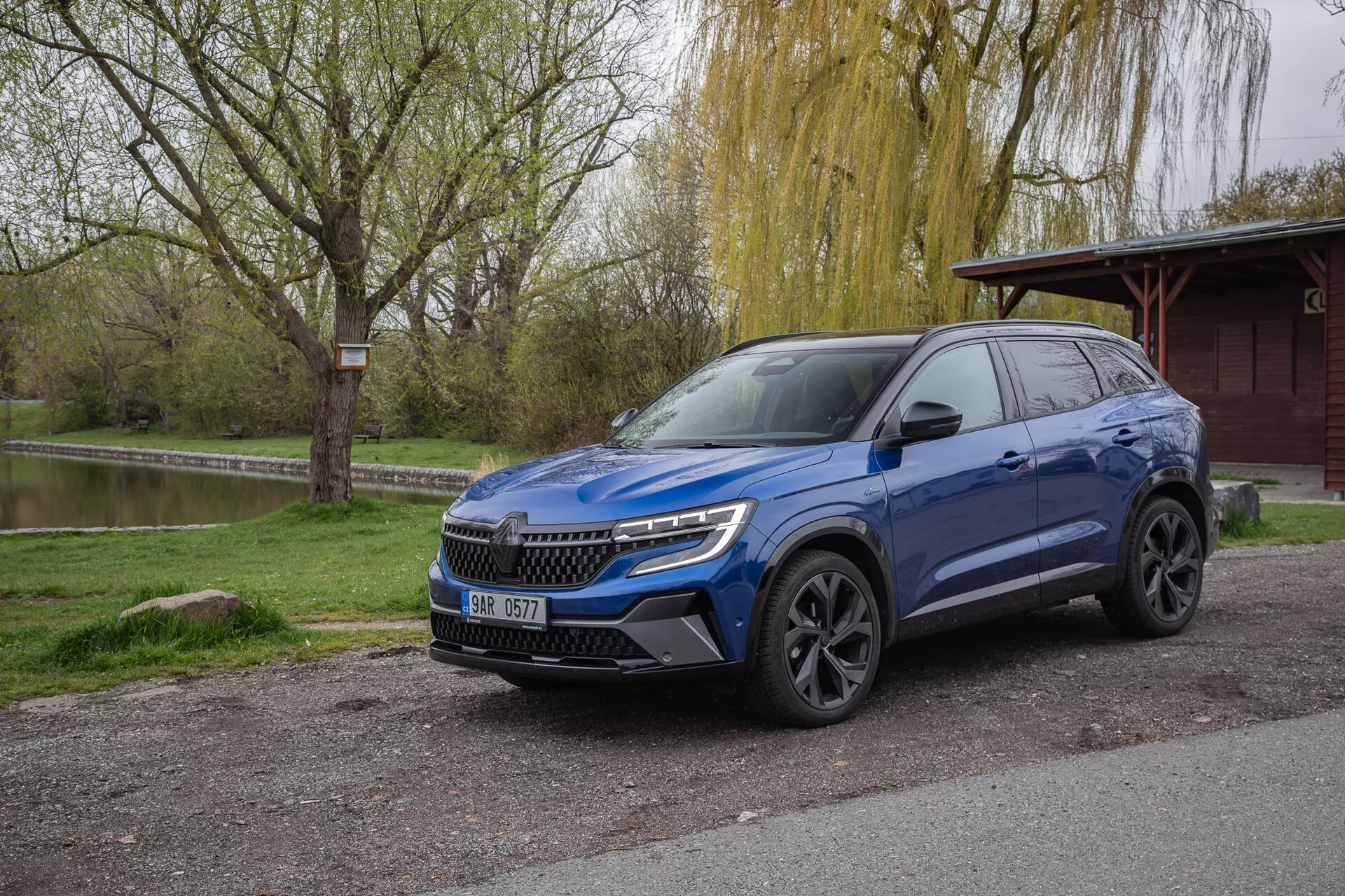 Crossover SUV Renault Austral 2022 v metalickej modrej farbe, zaparkované vonku ukazujúce čelný a bočný profil s LED svetlometmi a strešnými nosičmi, obklopené prírodným parkovým prostredím