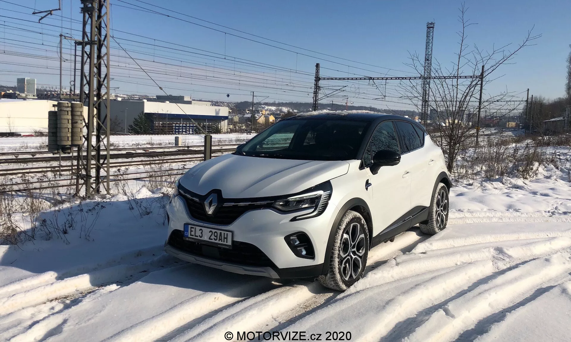 Obrázok zobrazuje biely Renault Captur 2020, kompaktné SUV zaparkované na zasneženej krajnici, zachytené z trojštvrťového predného uhla. Vozidlo sa vyznačuje charakteristickými LED dennými svetlami v tvare písmena C, projekčnými svetlometmi, prednou maskou s čiernymi akcentmi a výrazným logom Renault a viacramennými zliatinovými kolesami. Pozoruhodné dizajnové prvky zahŕňajú plastové opláštenie pozdĺž spodných hrán a okolo blatníkov. Prostredie naznačuje chladný zimný deň s priemyselným alebo polomestským pozadím.