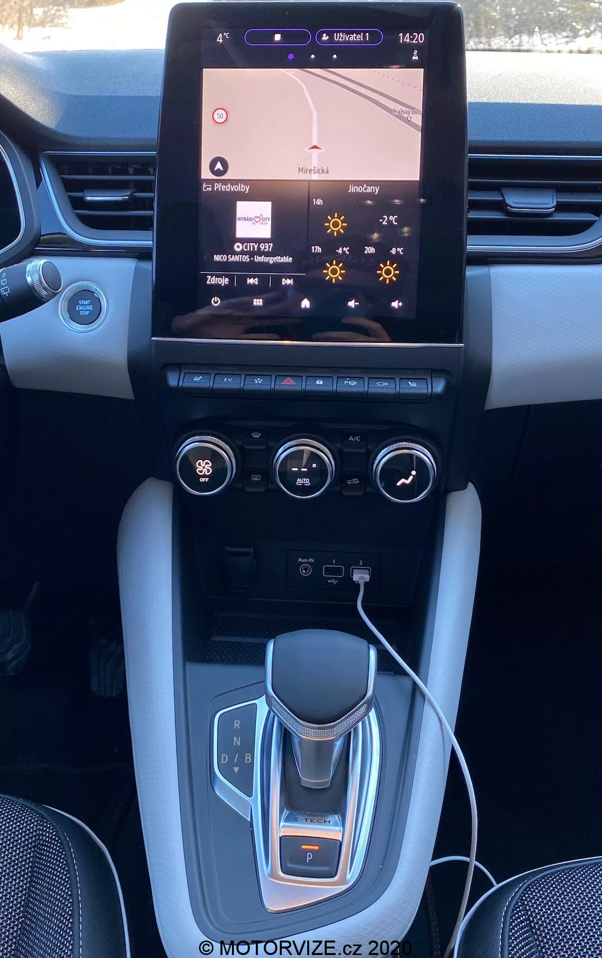 Interiér vozidla Renault Captur 2020 zobrazujúci stredovú konzolu a palubnú dosku s veľkým dotykovým infotainment systémom zobrazujúcim navigačnú mapu, dvojzónové klimatizačné ovládania, elektronický volič prevodovky, držiaky na nápoje, kefované kovové akcenty a možné luxusné povrchové úpravy.