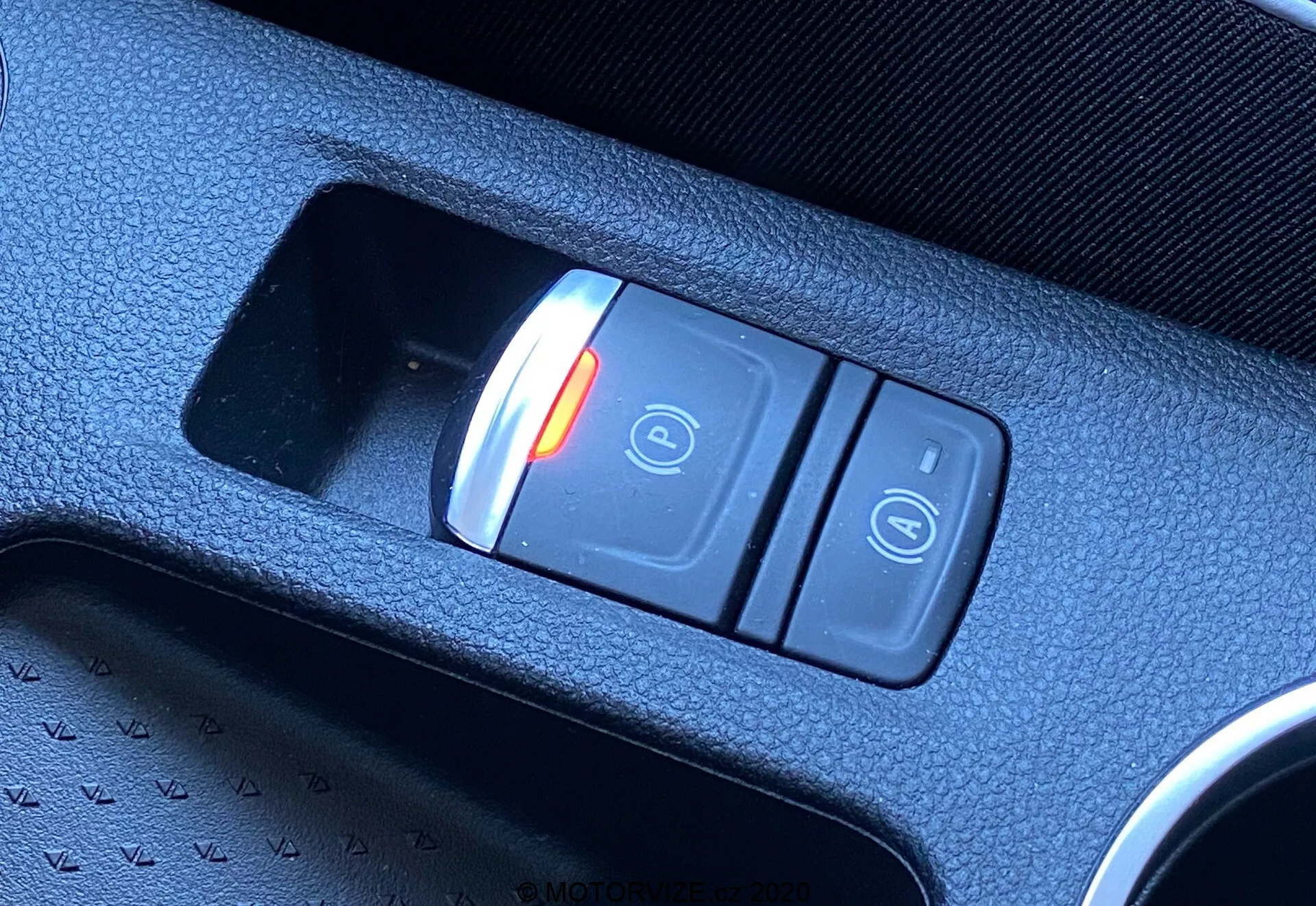Blízky diagonálny záber na elektronickú parkovaciu brzdu v interiéri Renault Captur 2020. Spínač umiestnený v centrálnej konzole obsahuje dve tlačidlá: osvetlené červené tlačidlo so symbolom zatvoreného zámku a 'P', čo naznačuje aktivovaný stav, a ďalšie tlačidlo so symbolom otvoreného zámku. Spínač je osadený v čiernej textúrovanej plastovej konzole, čo zdôrazňuje modernú technológiu vozidla.