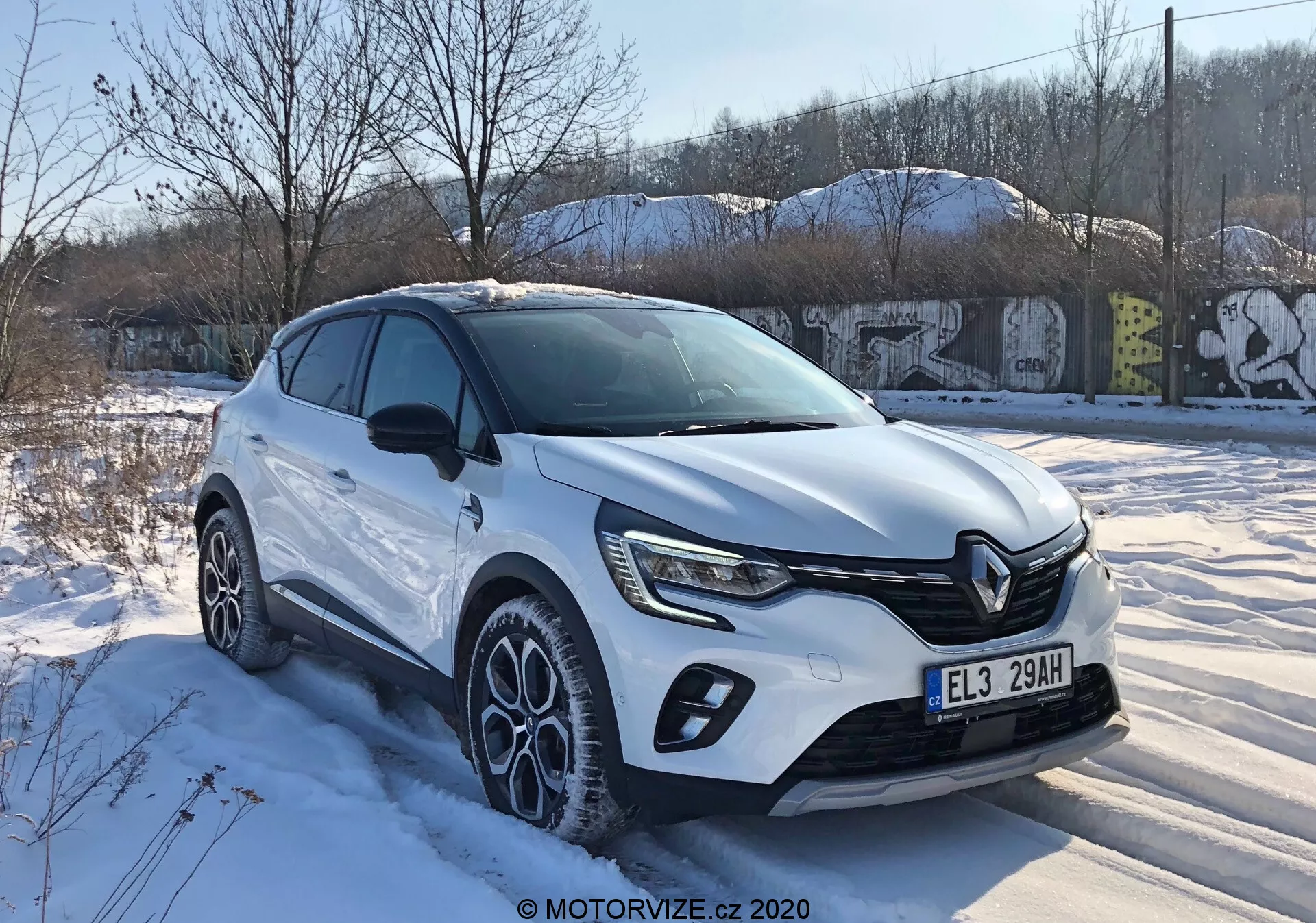 Renault Captur 2020 v trojštvrťovom prednom pohľade, ktorý zobrazuje moderný dizajn crossoveru s výraznou prednou mriežkou, ostrými LED svetlami, ochrannými lištami na karosérii, veľkými viacramennými zliatinovými kolesami a bielou karosériou za zimných snehových podmienok v mestskom prostredí.