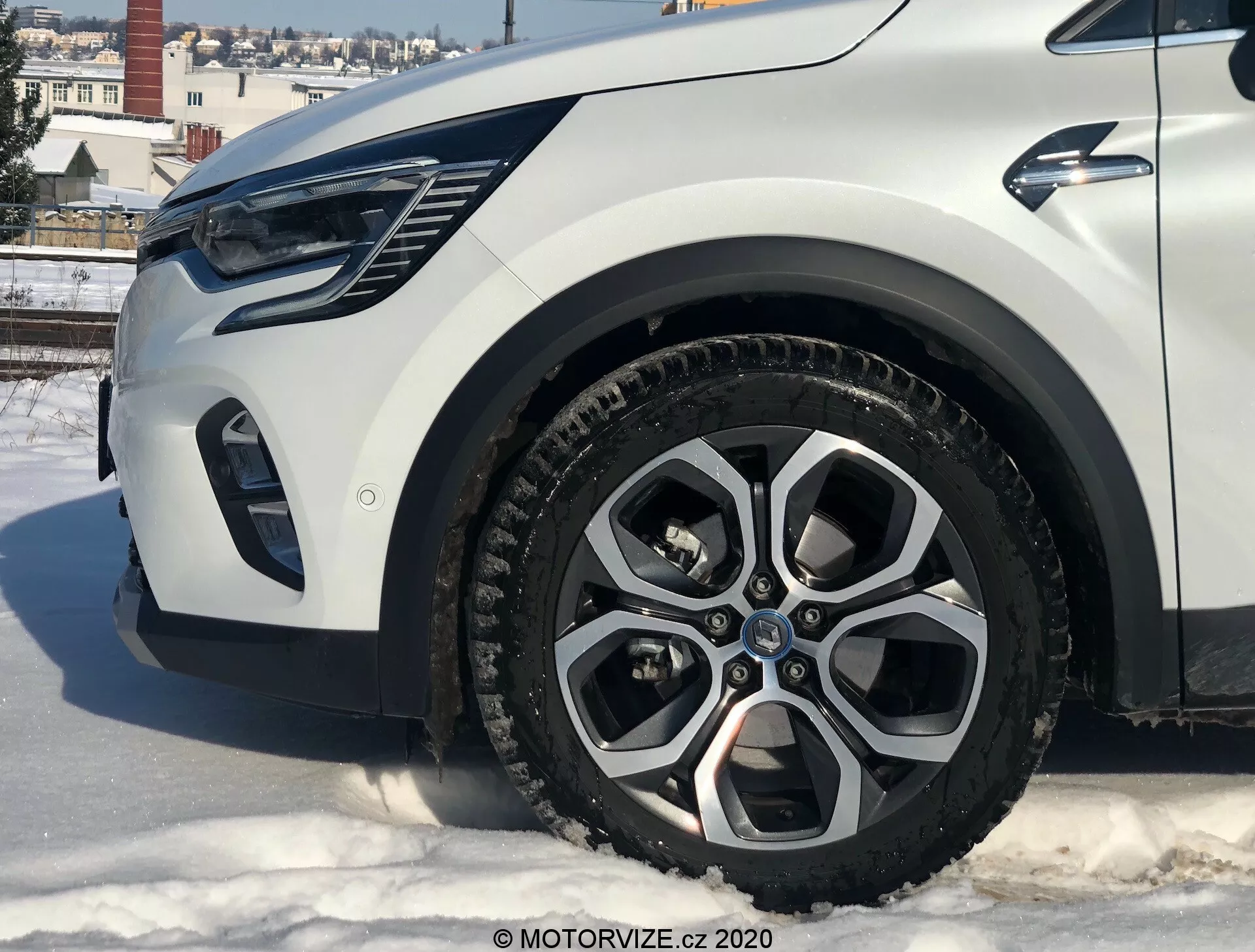 Detailný pohľad na Renault Captur 2020 z nízkeho uhla, ktorý ukazuje ľavú prednú časť, so zameraním na zliatinové koleso, blatník, nárazník a oblasť svetlometu. Auto sa nachádza na zasneženom povrchu v mestskom prostredí s budovami a jasnou oblohou.
