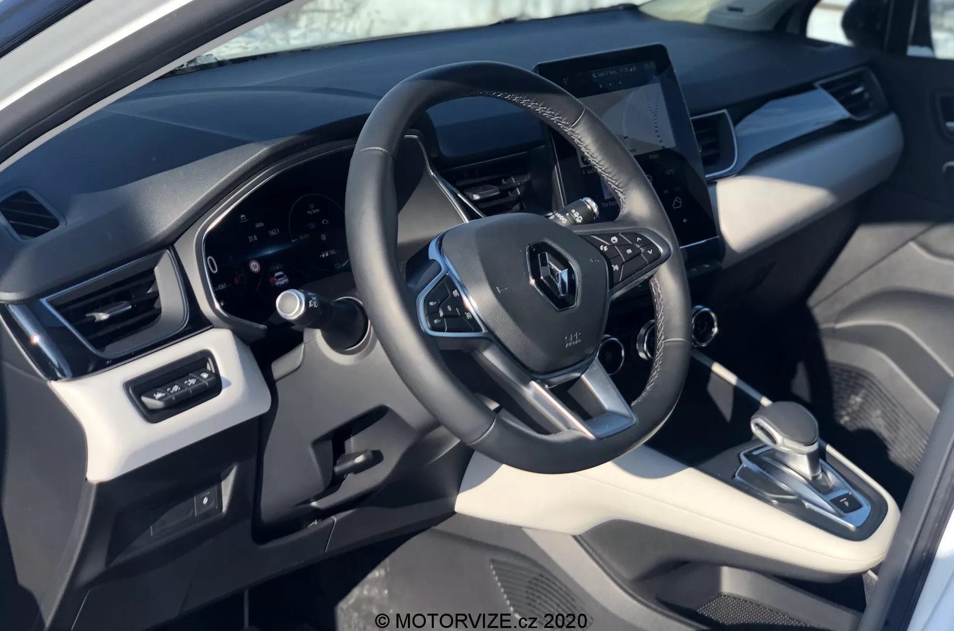 Interiér vozidla Renault Captur 2020 z pohľadu vodiča, ukazujúci volant s logom Renault, namontované ovládacie prvky, digitálny prístrojový panel za ním a stredne umiestnený dotykový infotainment systém. Snímka zdôrazňuje zmes syntetickej kože a plastových materiálov s kovovými akcentmi, automatickou prevodovkou, systémom klimatizácie a nastavením okien a zrkadiel.