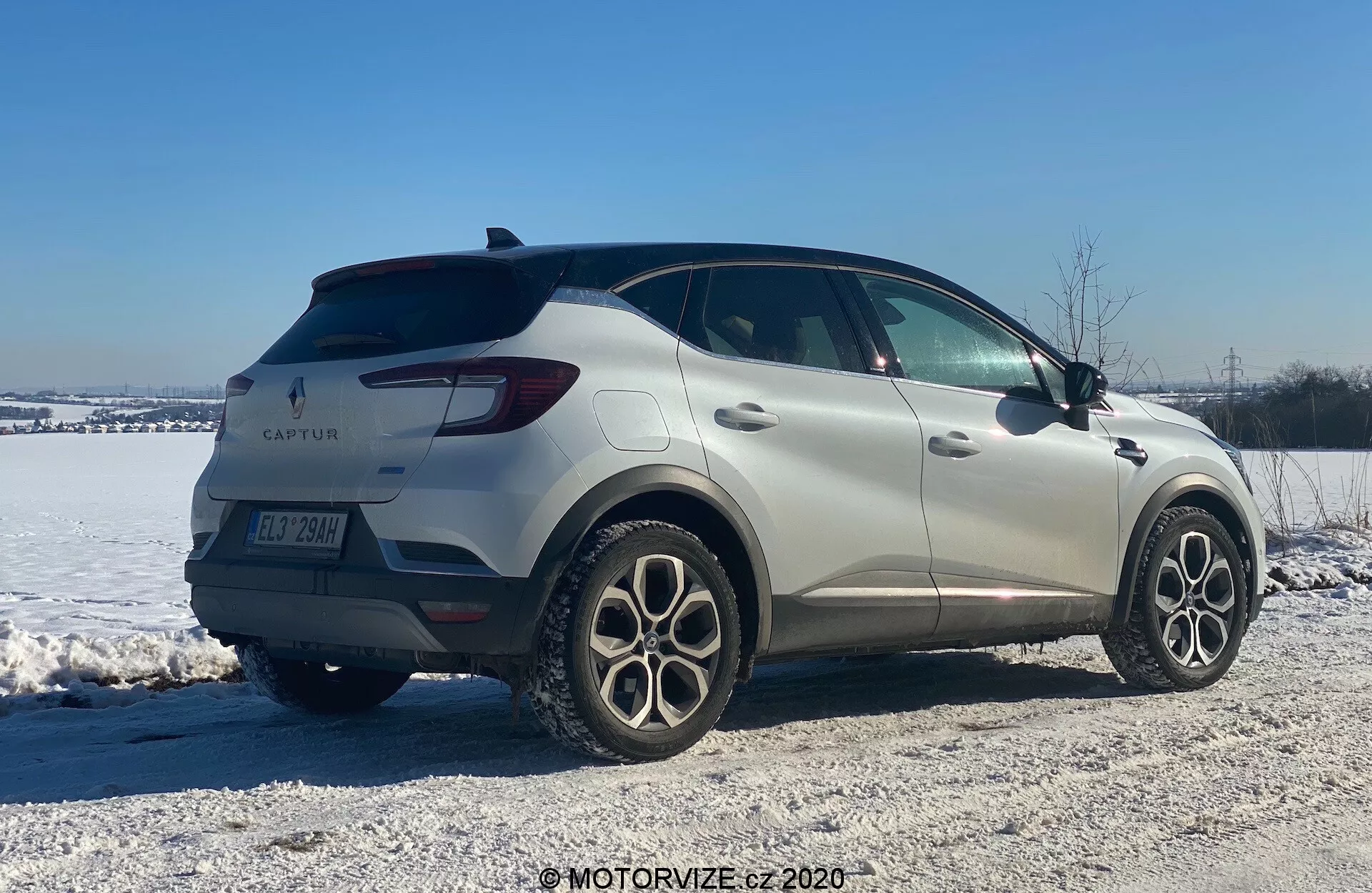 Obrázok Renaultu Captur 2020, kompaktného SUV, v zimnom prostredí. Zobrazený zo zadného trojštvrťového pohľadu, vozidlo vystavuje elegantný dizajn s hladkými líniami a LED zadnými svetlami. Viditeľné sú znaky výrobcu a názov modelu spolu s viaclúčovými zliatinovými kolesami, zadnými parkovacími senzormi a bočnými zrkadlami s integrovanými smerovými svetlami. Umiesnené na zasneženej štrkovej ploche, obraz naznačuje schopnosť jazdy v rôznorodom teréne a zimných podmienkach.
