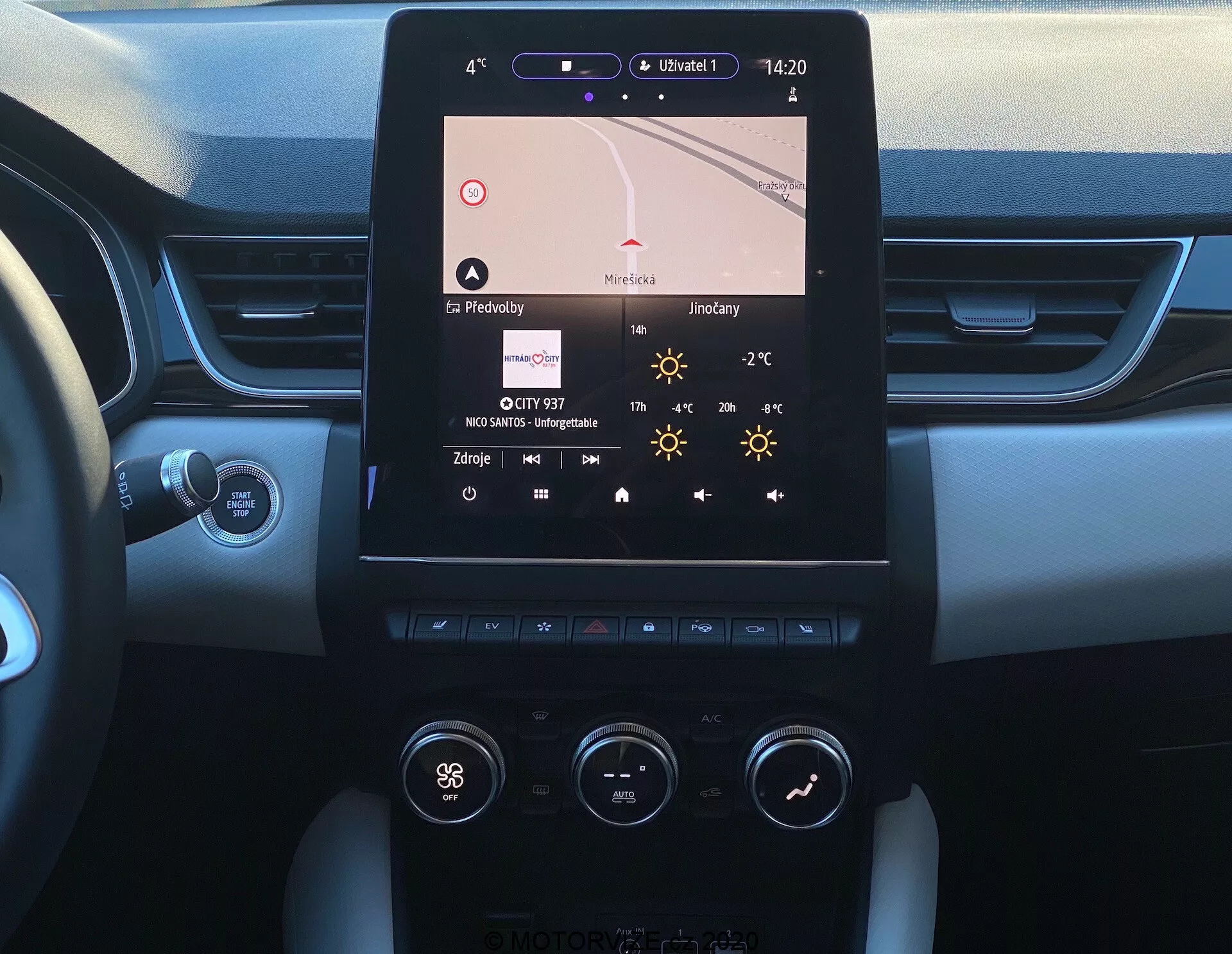 Obrázok zobrazuje stredovú konzolu Renault Captur 2020 z pohľadu sedadla vodiča. Hlavným prvkom je vertikálne orientovaný dotykový displej infotainmentu zobrazujúci navigáciu, čas (14:20), teplotu (27°C) a podrobnosti o rádiu. Nižšie je viditeľný dvojzónový systém klimatizácie s gombíkmi a tlačidlami naznačujúcimi nastavenia ako 'Hi' pre teplotu na strane vodiča. Konzola kombinuje kovové a plastové materiály okolo obrazovky a ovládacích prvkov. Okraje prístrojovej dosky odhaľujú mäkký dvojtónový dizajn a čiastočne viditeľné je tlačidlo Start/Stop. Viditeľný je aj stĺpik riadenia s ovládacou pákou, pravdepodobne pre smerovky alebo iné funkcie.
