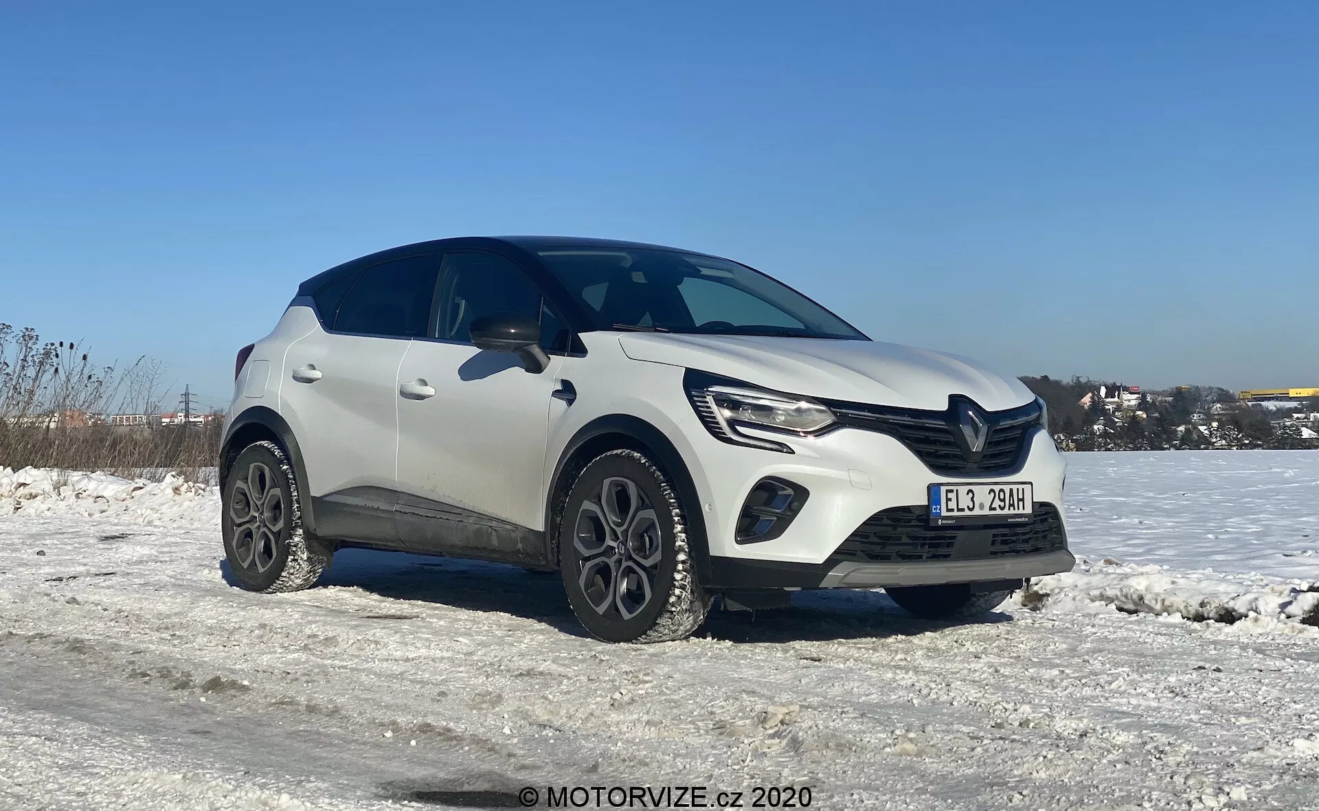 Trojštvrťový pohľad na biely kompaktný crossover SUV Renault Captur 2020 v zimnom prostredí, so zasneženým povrchom a jasnou modrou oblohou. Obraz zvýrazňuje ľavú prednú stranu vozidla a časť jeho bočného profilu, urobený z mierne nižšieho uhla zdôrazňujúceho kolesá a dolnú polovicu vozidla. Moderný dizajn auta zobrazuje výrazné línie na kapote, elegantné LED svetlomety s dennými svetlami a výraznú prednú masku. Unikátne zliatinové kolesá a plastové obklady okolo oblúkov kolies pridávajú vozidlu robustný vzhľad. Tiene pod autom naznačujú, že fotografia bola urobená na poludnie alebo skoro popoludní za denného svetla.