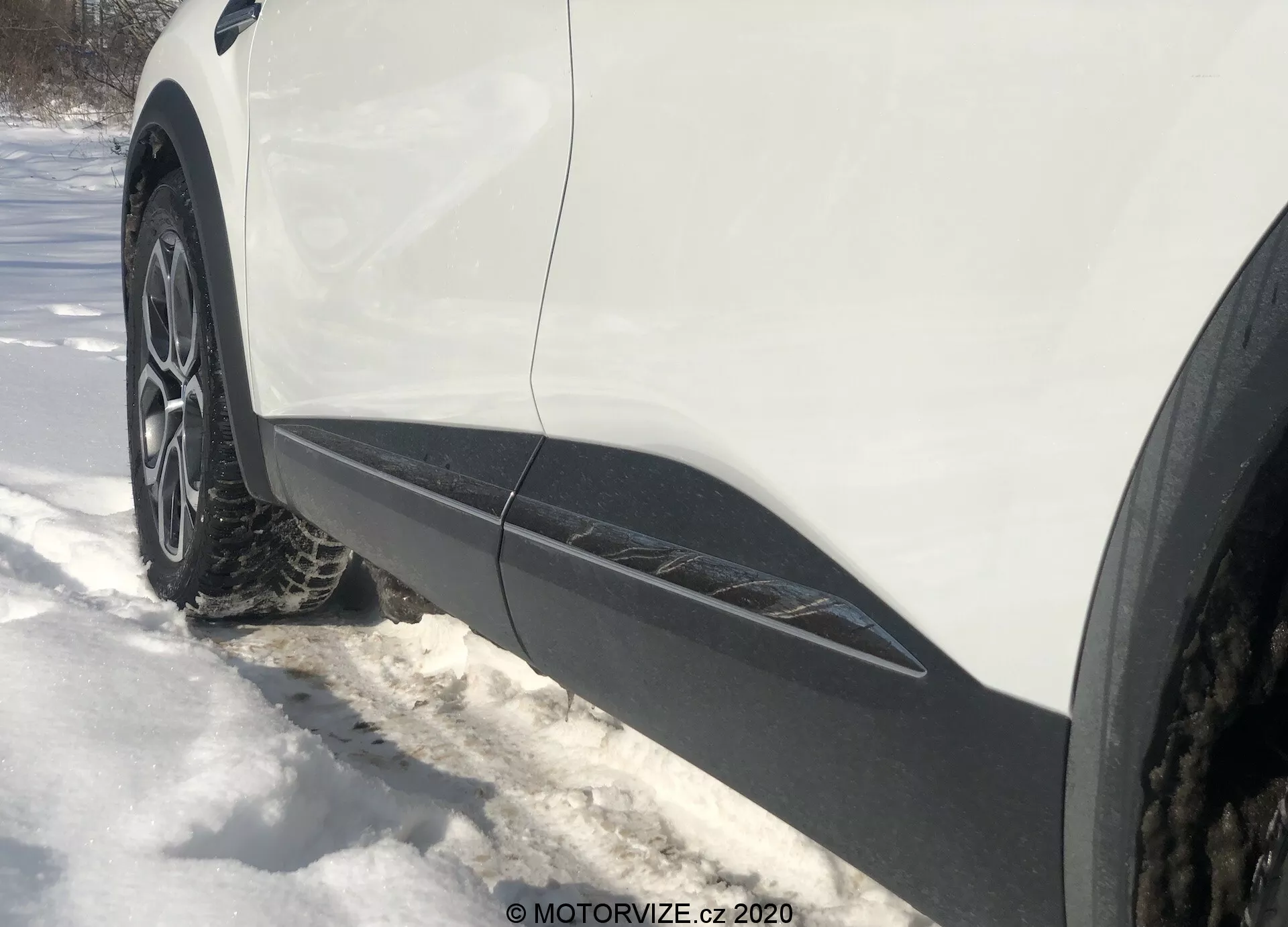 Blízky, šikmý bočný pohľad z úrovne zeme na predné koleso a blatník zo strany vodiča Renault Captur 2020. Viacramenné hliníkové koleso so zimnou alebo celoročnou pneumatikou je obklopené snehom. Biely karosériový diel s čiernym obložením vedie pozdĺž spodnej hrany, čo naznačuje crossover alebo SUV. Osvetlenie naznačuje slnečný deň s viditeľnými tieňmi.