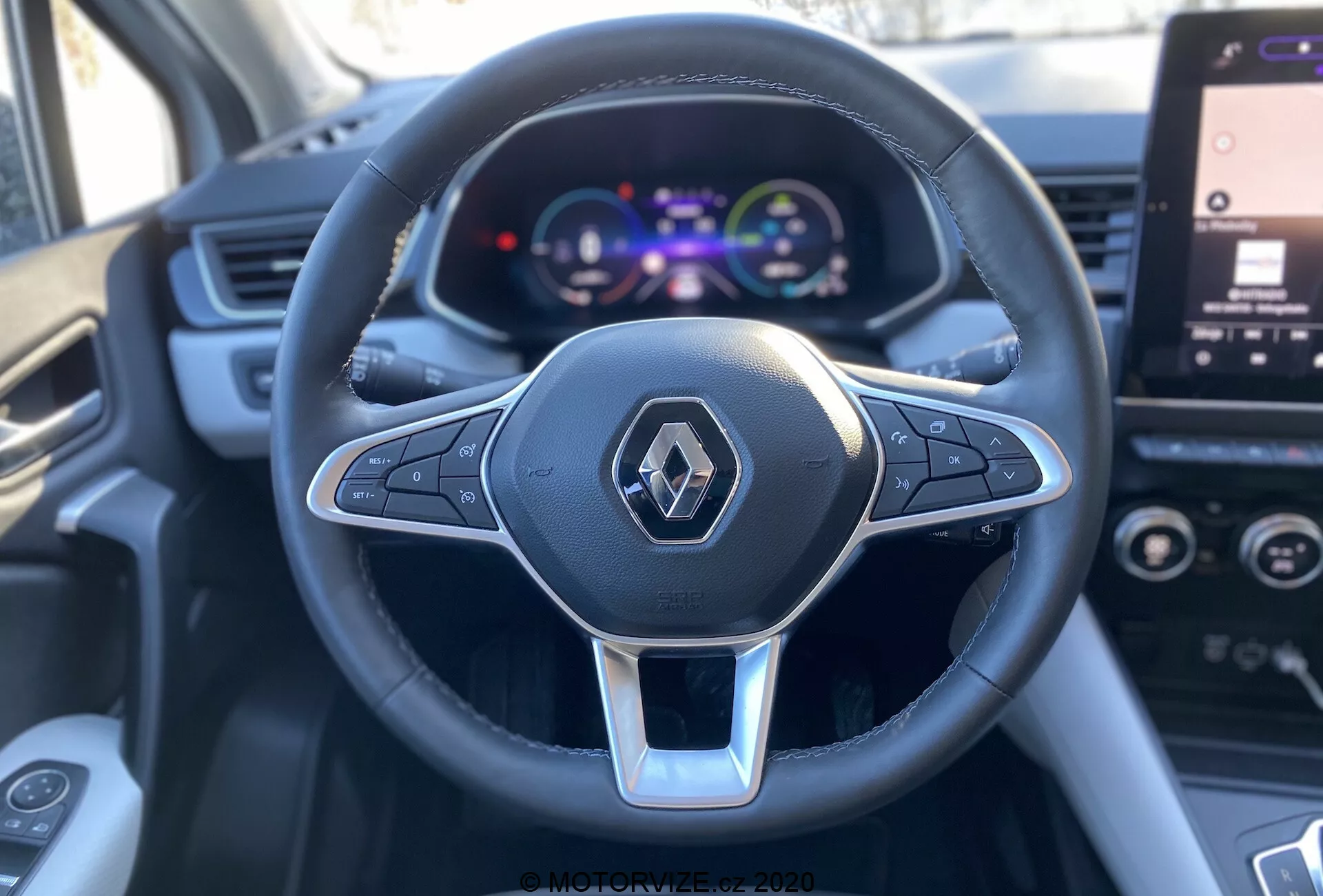 Detailný pohľad z prvej osoby zo sedadla vodiča Renault Captur 2020, zameraný na multifunkčný volant s troma ramenami, potiahnutý kožou so strieborným akcentom a logom Renault, spolu s farebným digitálnym prístrojovým panelom zobrazujúcim rýchlomer a detaily vozidla, s čiastočne viditeľnou dotykovou obrazovkou infotainmentu v pozadí.