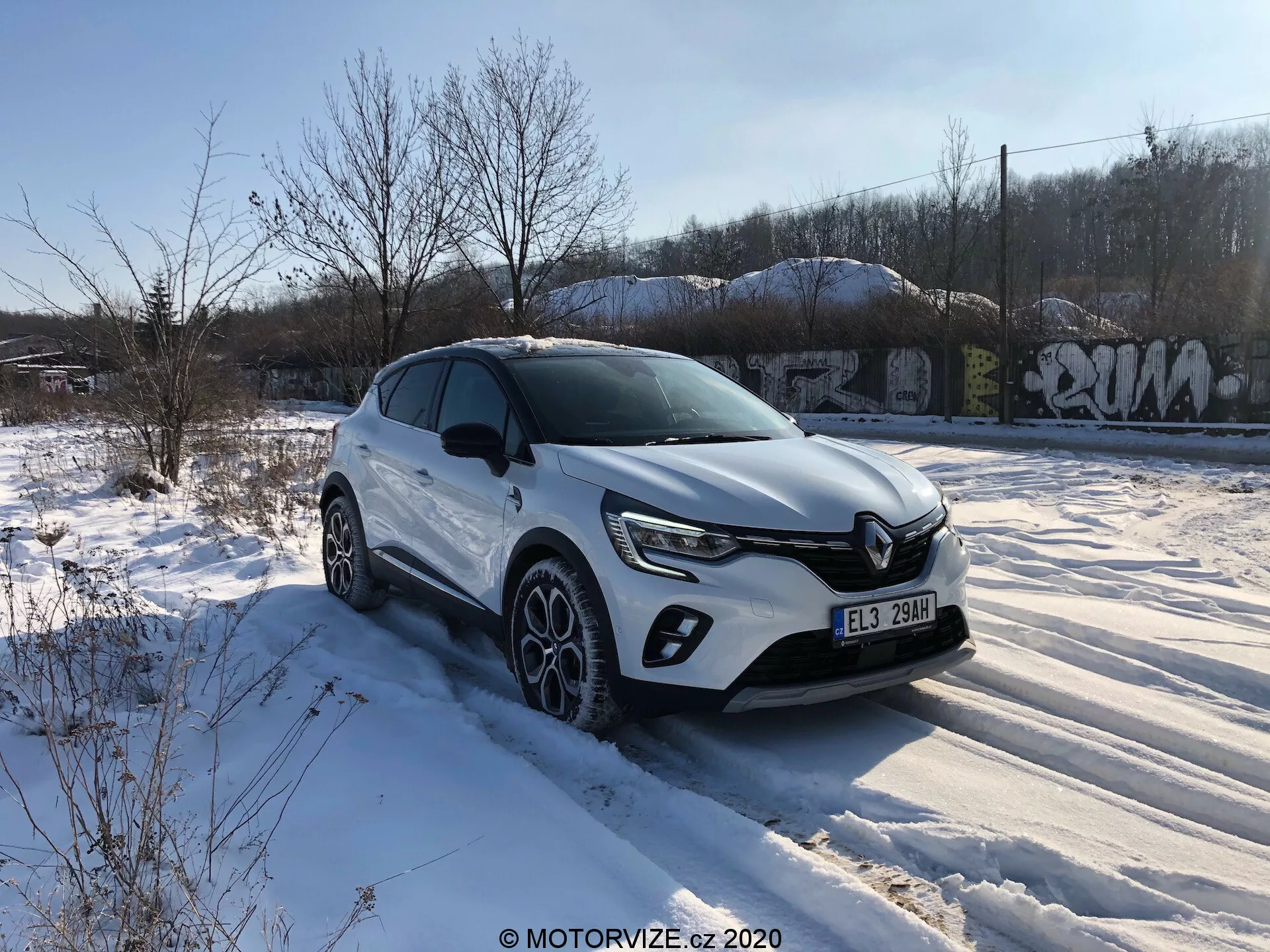 Biely crossover SUV Renault Captur 2020 je zaparkovaný na zasneženom teréne s vegetáciou a graffiti pokrytými štruktúrami v pozadí, pod prevažne jasnou oblohou s niekoľkými oblakmi. Obrázok bol nasnímaný z trištvrťového predného uhla zameraného na stranu vodiča, ukazujúce prednú masku s chrómovým emblémom a modernými LED svetlometmi. SUV má predné LED denné svietenie, kryty hmlových svetiel, charakterové línie na kapote, plastové obloženie okolo blatníkov, integrované smerovky v bočných zrkadlách, multifunkčné zliatinové kolesá so zimnými pneumatikami a silnú ramennú líniu. Zvýšená pozícia naznačuje vyššiu svetlú výšku, ktorá je vhodná pre rôzne terény.