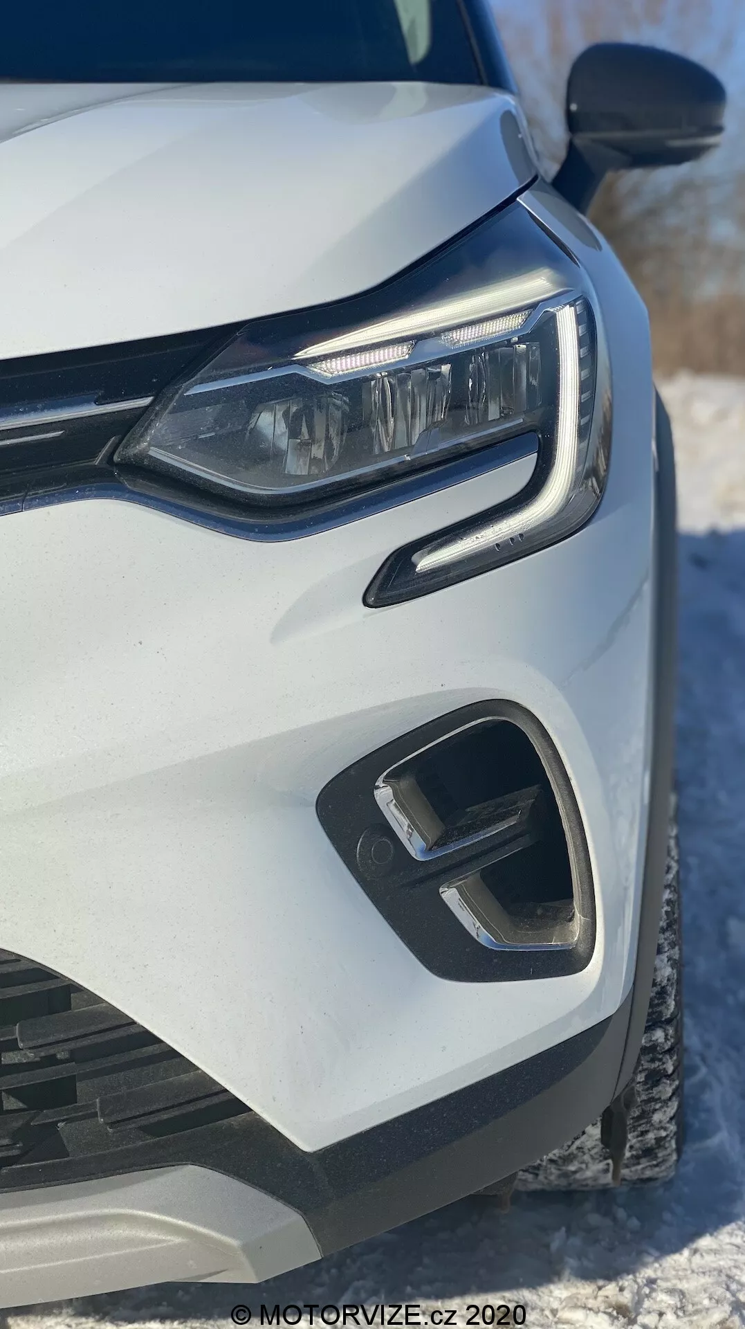 Detailný záber prednej ľavej strany bieleho Renault Captur 2020, zameraný na moderný LED svetlomet, geometrický predný nárazník a prvky mriežky, zachytený z nízkeho čelného uhla so snehom v pozadí