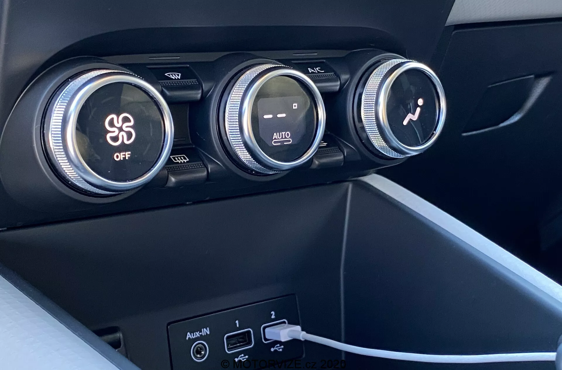 Zblízka interiér Renault Captur 2020 so zameraním na klimatizačný systém. Na obrázku sú tri veľké ovládacie gombíky na prístrojovej doske. Prvý gombík ovláda klimatizáciu, označený symbolom snehovej vločky a tlačidlom 'OFF'. Stredný gombík reguluje teplotu s červenými a modrými prechodmi farieb a indikáciou 'AUTO'. Tretí gombík s ikonou ventilátora nastavuje rýchlosť ventilátora. Pod gombíkmi sú dva USB porty a pomocný vstup, s káblom zapojeným do jedného z USB portov. Materiály sú matné, v tmavom plaste alebo syntetických materiáloch, s moderným minimalistickým dizajnom.