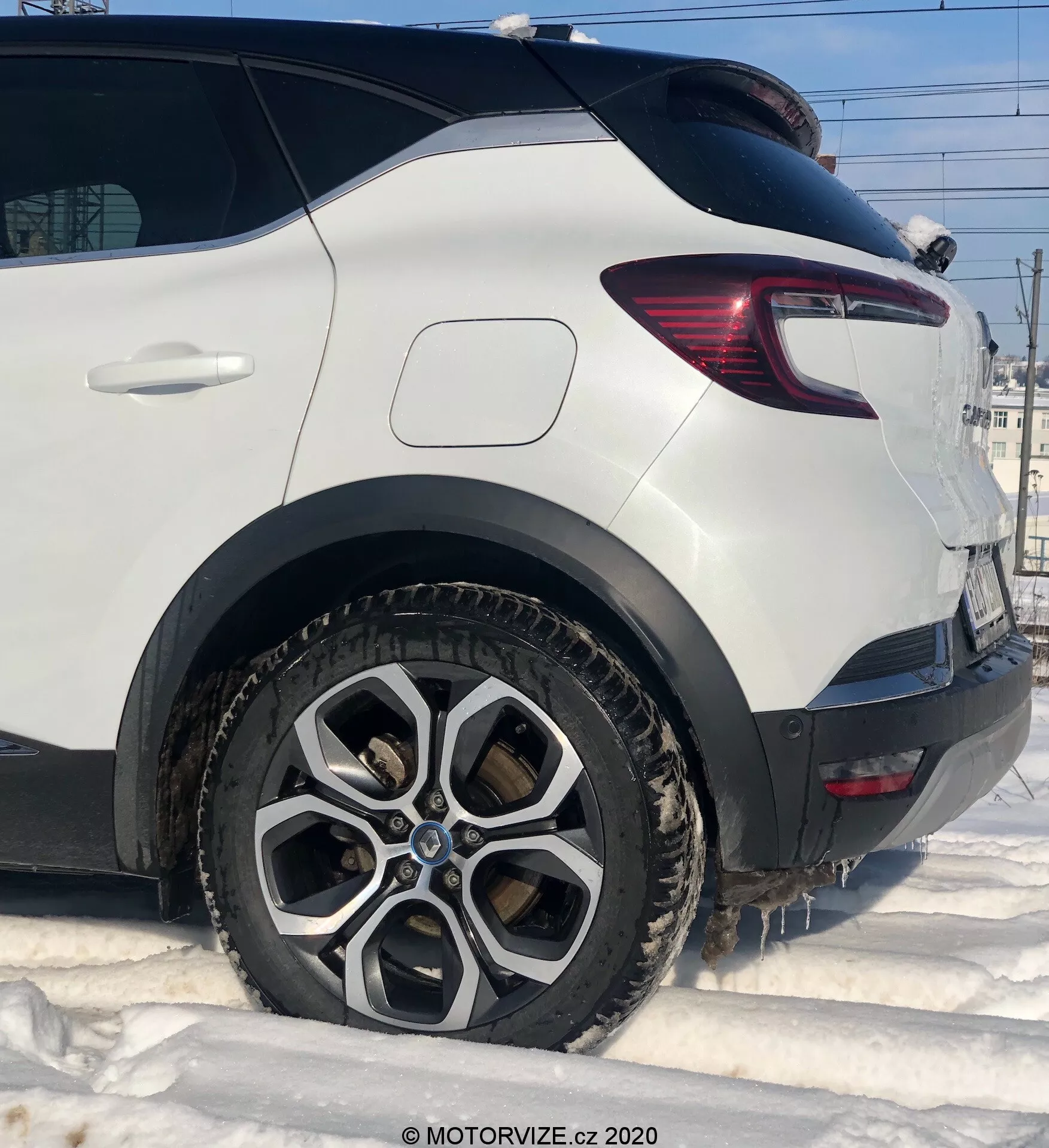 Detailný záber na biely Renault Captur 2020, ukazujúci zadnú stranu spolujazdca z nízkeho uhla. Zvýraznené sú zadné koleso s veľkolepým viacpaprskovým dizajnom, špinavé od blata a snehu, naznačujúce zimné podmienky. Obrázok zachytáva čistý zadný blatník nad oblúkom kolesa, moderné zadné svetlo s červenými a čírymi LED segmentmi a robustné plastové opláštenie za kolesom. Šikmé okno na zadnom hatchu prispieva k aerodynamickému tvaru vozidla.