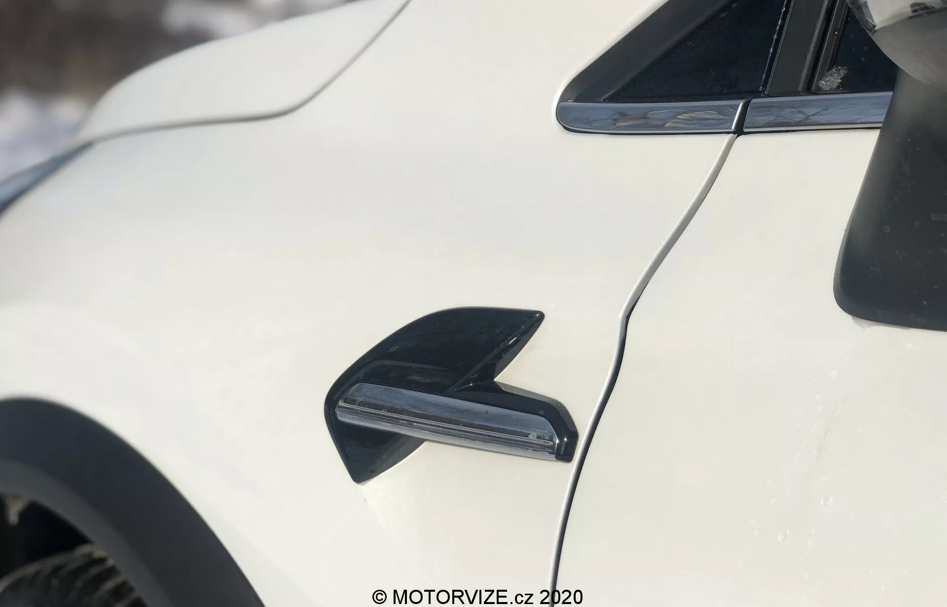 Detailný záber na exteriér Renault Captur 2020, zameriavajúci sa na bok bieleho vozidla. Obrázok obsahuje čiernu, aerodynamicky tvarovanú anténu v tvare žraločej plutvy v ľavom hornom rohu, určenú na príjem rádia. Pod anténou je zvýraznený tvarovaný čierny vetrací otvor alebo prívod vzduchu, pravdepodobne na chladenie bŕzd alebo zlepšenie prúdenia vzduchu. Aerodynamická línia strechy vozidla sa jemne kloní smerom k zadnej časti, s lesklým bielym lakom odrážajúcim svetlo. Rozmazané pozadie naznačuje zasnežené vonkajšie prostredie.