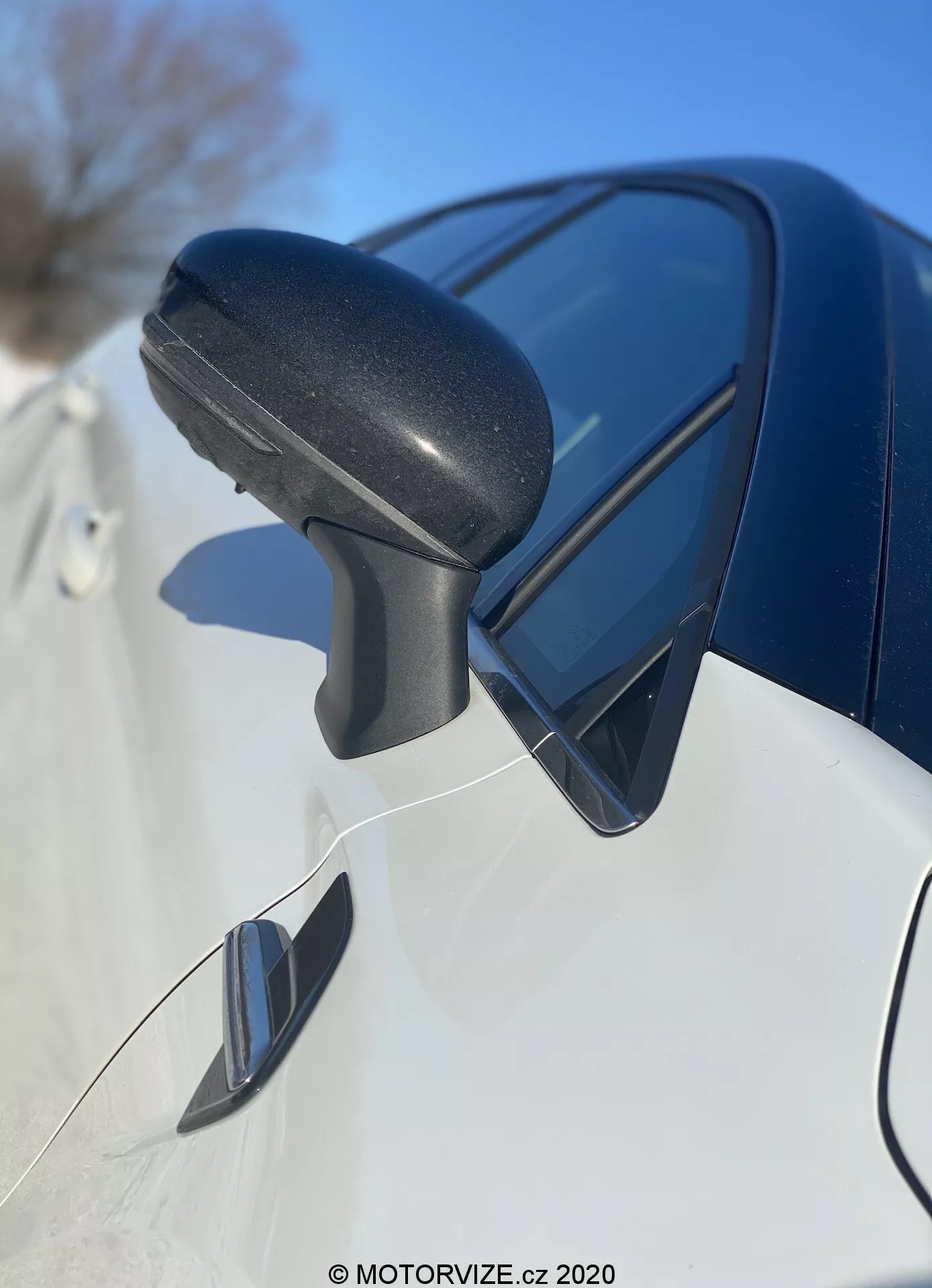 Detailný pohľad na bočné zrkadlo Renault Captur 2020 zpredu dozadu, zamerať na čierny textúrovaný plastový kryt zrkadla s integrovaným smerovým svetlom a odrážajúcim modrú oblohu. Elegantný tmavý farbený panel karosérie pod ním a potenciálne zrkadlo slepého uhla naznačujú moderný dizajn a pokročilé funkcie.
