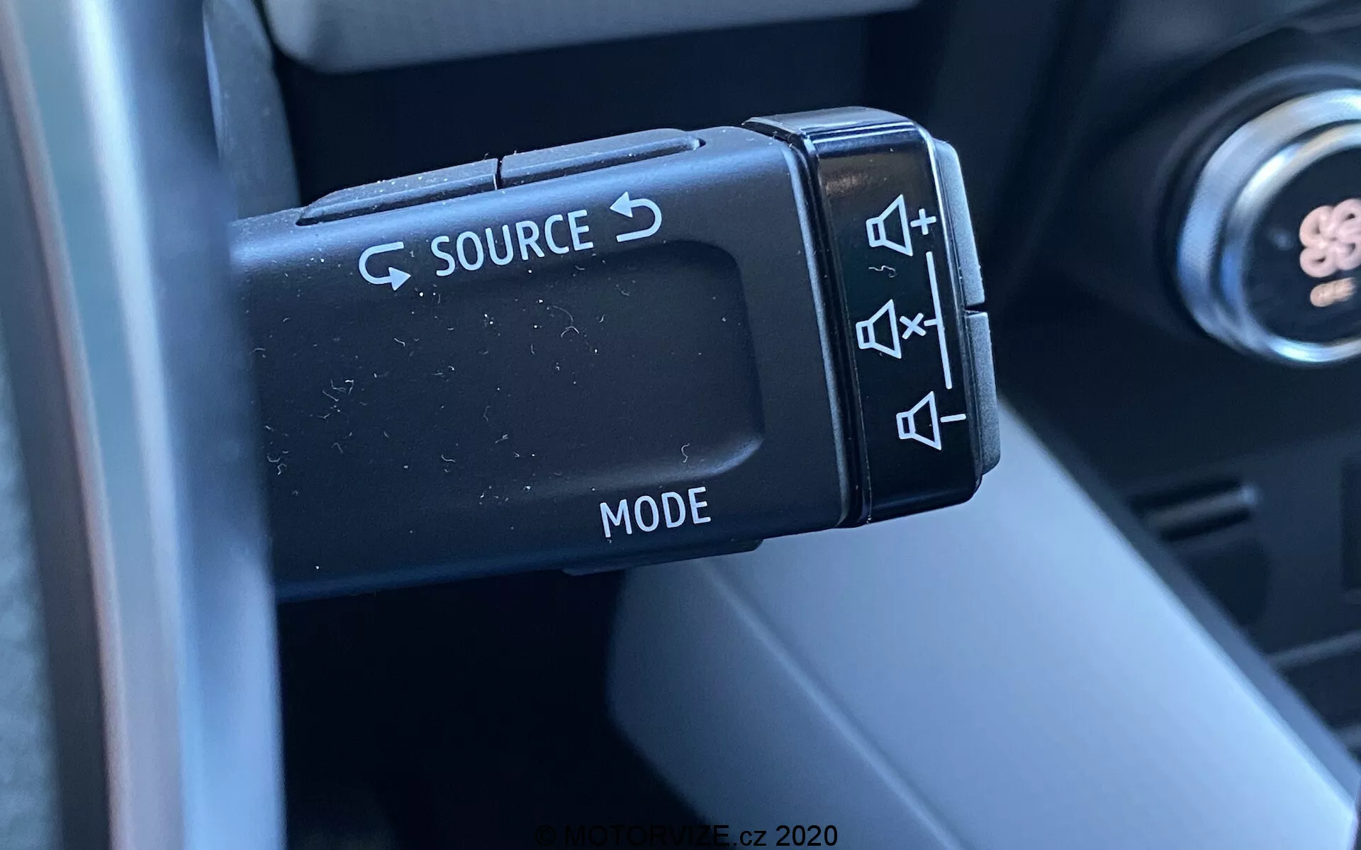 Detailný záber na ovládanie audia na volante vo vozidle Renault Captur 2020, sústreďujúci sa na ovládaciu páčku na ľavej strane s tlačidlami na zmenu zdroja (SOURCE), nastavenie hlasitosti, preskočenie skladby a výber režimu (MODE). Fotografia zachytáva detaily matne dokončených plastových tlačidiel a ich ergonomický dizajn, pričom časť stĺpika riadenia a interiéru je v pozadí rozostrená, zdôrazňujúca funkčnosť a pohodlie pre vodiča.