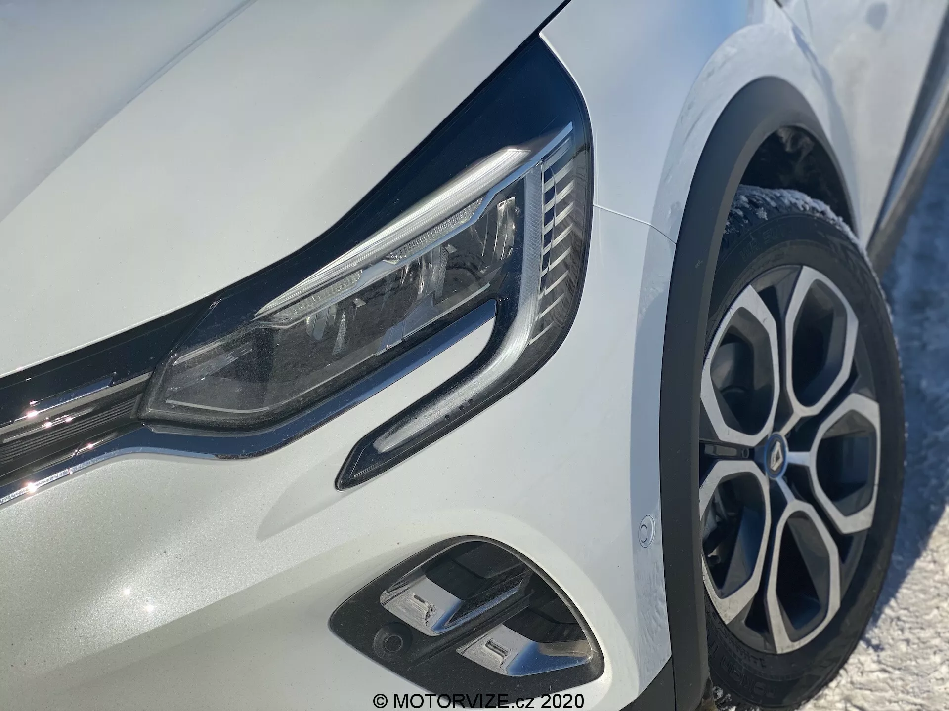Detailný záber na Renault Captur 2020 zameraný na prednú ľavú časť, ukazujúci elegantné LED reflektory, štýlový viacramenný zliatinový disk, dynamický predný nárazník so vzduchovými otvoreniami a lesklé chrómové akcenty na jasne bielom karosérii.