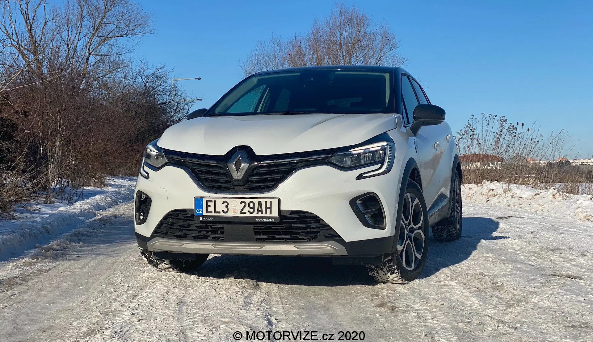 Čelný pohľad na biely Renault Captur 2020, fotografovaný pod uhlom mierne zvýrazňujúcim ľavý bok. Moderné vozidlo predstavuje charakteristický predný mriežkový dizajn Renault s veľkým centrálnym logom, ostrými uhlovými líniami a tvarovanými kontúrami. Zobrazené sú celá predná mriežka, LED svetlomety a uhlové denné svietenie. Športový nárazník má výrazné bočné vzduchové vstupy a stredovú časť pravdepodobne slúžiacu na chladenie. Kapota má zvýšené línie pre svalnatý vzhľad. Časť predných kolies a viaclúčové hliníkové ráfiky sú viditeľné na zasneženej ploche, čo naznačuje zimné podmienky. Európska poznávacia značka a lesklý lak odrážajúci slnečné lúče naznačujú slnečný, jasný deň. Pokročilé systémy asistencie vodiča, LED osvetlenie a hybridný/elektrický pohon sú pravdepodobne odvodené z 'E' na poznávacej značke.