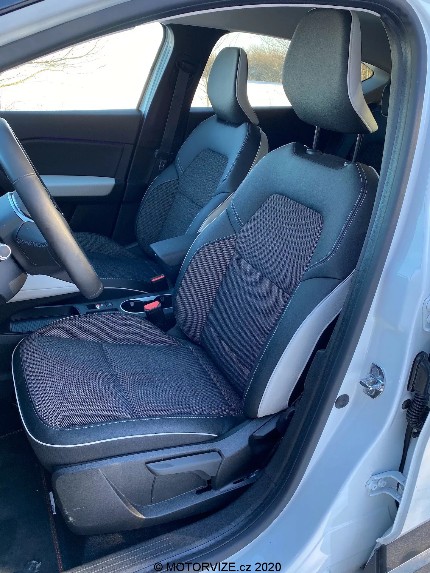 Pohľad na interiér Renault Captur 2020 z ľavej strany s otvorenými dverami na strane vodiča, ukazujúcim čierno-sivé látkové a kožené sedadlá, stredovú konzolu, volant a časť prístrojovej dosky.