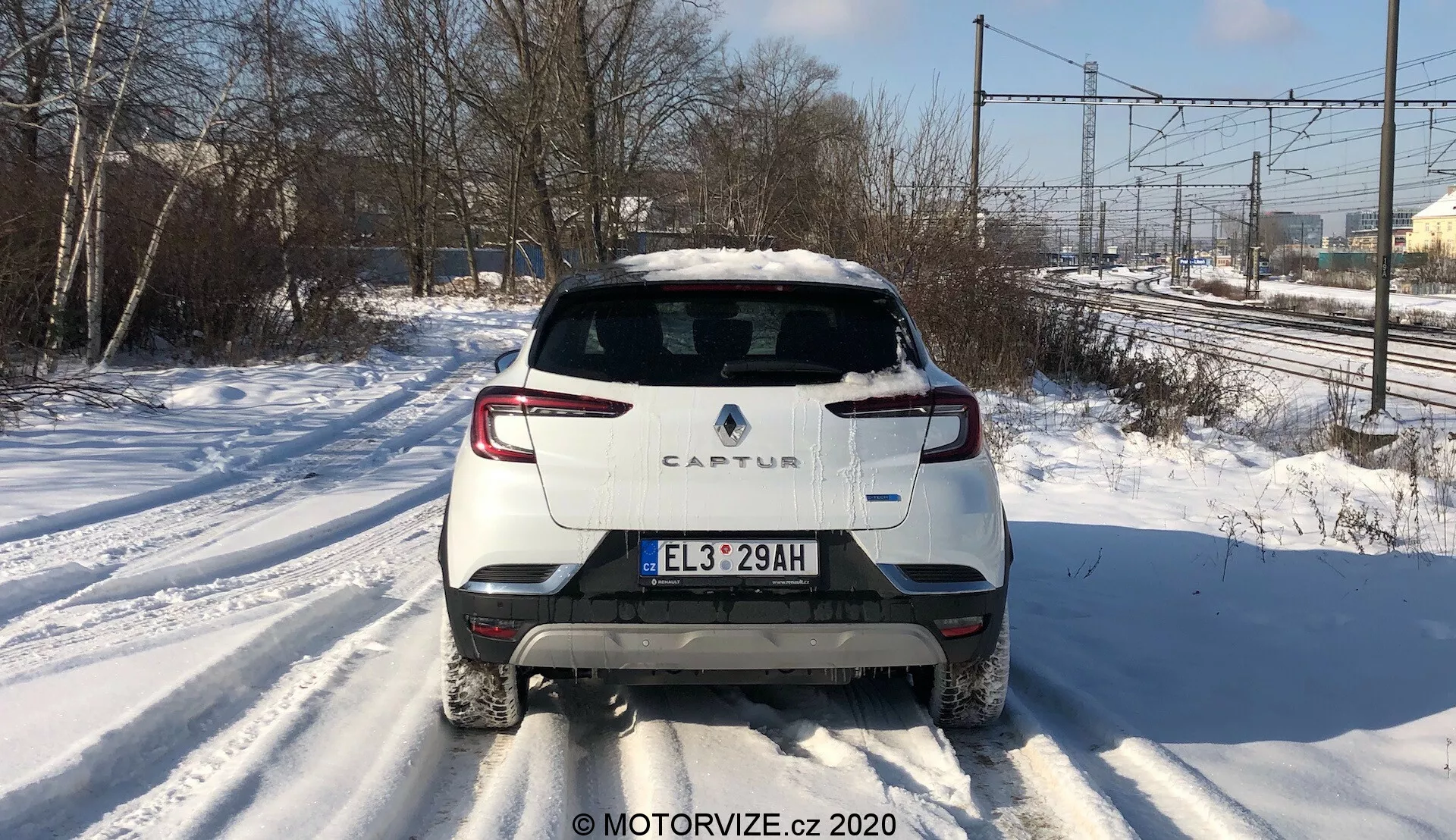 Pohľad zozadu na Renault Captur 2020 na zasneženej ceste. Obrázok zdôrazňuje výrazný zadný dizajn, vrátane dvojfarebnej schémy s bielym telom a kontrastnou strechou, moderného zadného LED osvetlenia, európskej poznávacej značky, zadného stierača a jedinej výfukovej rúry na pravej strane. Fotografia zachytáva elektrické vedenia a stromy v pozadí za denného svetla, čo naznačuje predmestský alebo polovenkovský kontext.