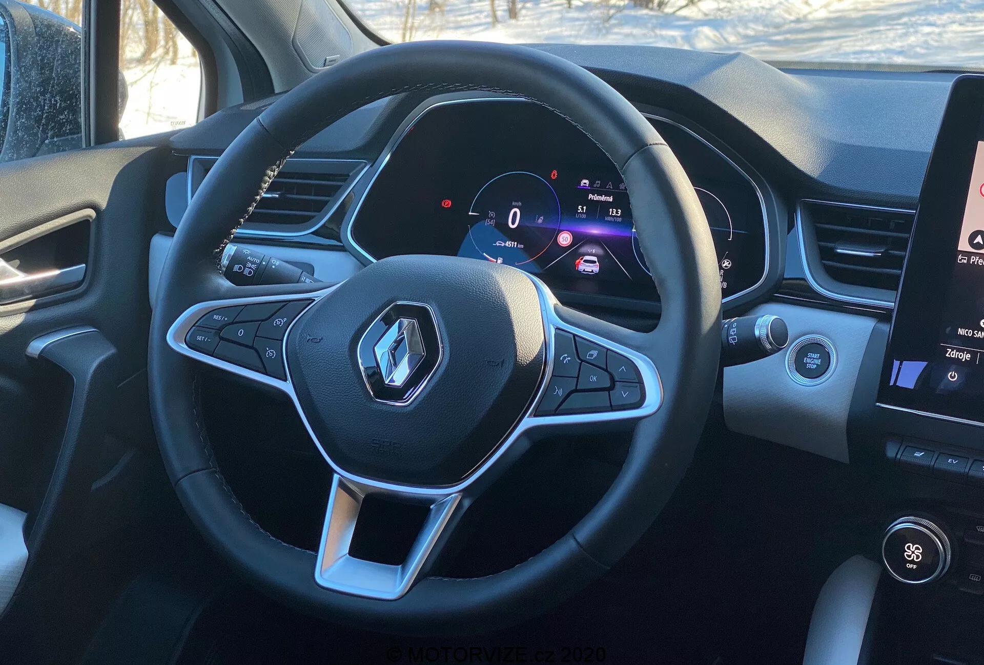 Interiér vozidla Renault Captur 2020 z pohľadu vodiča, zvýrazňujúci športový, plochý multifunkčný volant s logom Renault a ovládacími tlačidlami pre funkcie audia a tempomatu. Digitálny prístrojový panel zobrazuje stojacu rýchlosť 0 km/h vedľa otáčkomera, čo znamená modernú technológiu a prispôsobiteľné informácie pre vodiča. Na pravej strane je viditeľná časť vertikálneho dotykového infotainment systému, ktorý zobrazuje ovládanie teploty a mediálne informácie, za denných podmienok, ponúkajúci realistický pohľad z vodičovho sedadla.