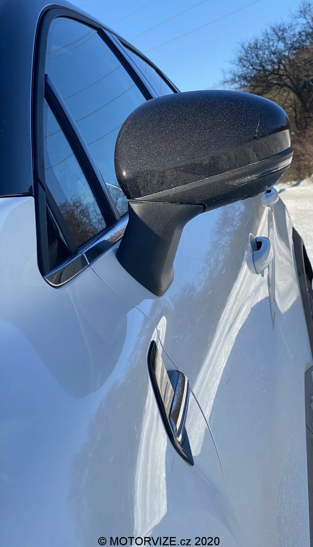 Detailný záber na bočné zrkadlo, dvere a predný štvrťpanel bieleho Renault Captur 2020. Fotografia, urobená z boku tesne pred bočným zrkadlom pod uhlom nahor, zvýrazňuje moderný, aerodynamicky účinný dizajn bočného zrkadla s čiernou textúrovanou spodnou časťou pre trvanlivosť. Prúdenkový držiak hladko prechádza do rámu dverí. Zapustená kľučka dverí a charakteristická línia, ktorá ňou prechádza, naznačujú športový, štíhly dizajn. Časť svetlometu a odraz jasnej oblohy v okne umocňujú toto dynamické prevedenie.