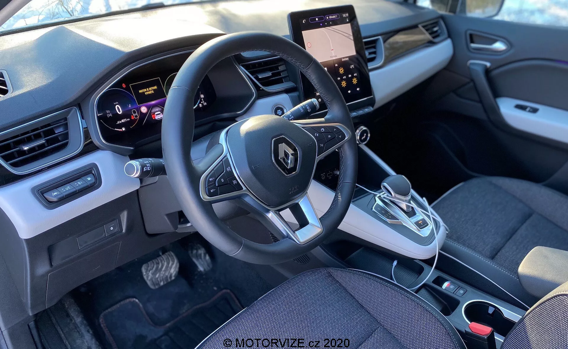 Pohľad na interiér Renault Captur 2020 zo zadného sedadla, zameraný na prednú kabínu, vrátane palubnej dosky, volantu s namontovanými ovládačmi, prístrojového panelu, stredovej konzoly s dotykovým displejom a ovládaním klimatizácie, automatickej radiacej páky a moderného mixu látkových a plastových prvkov.