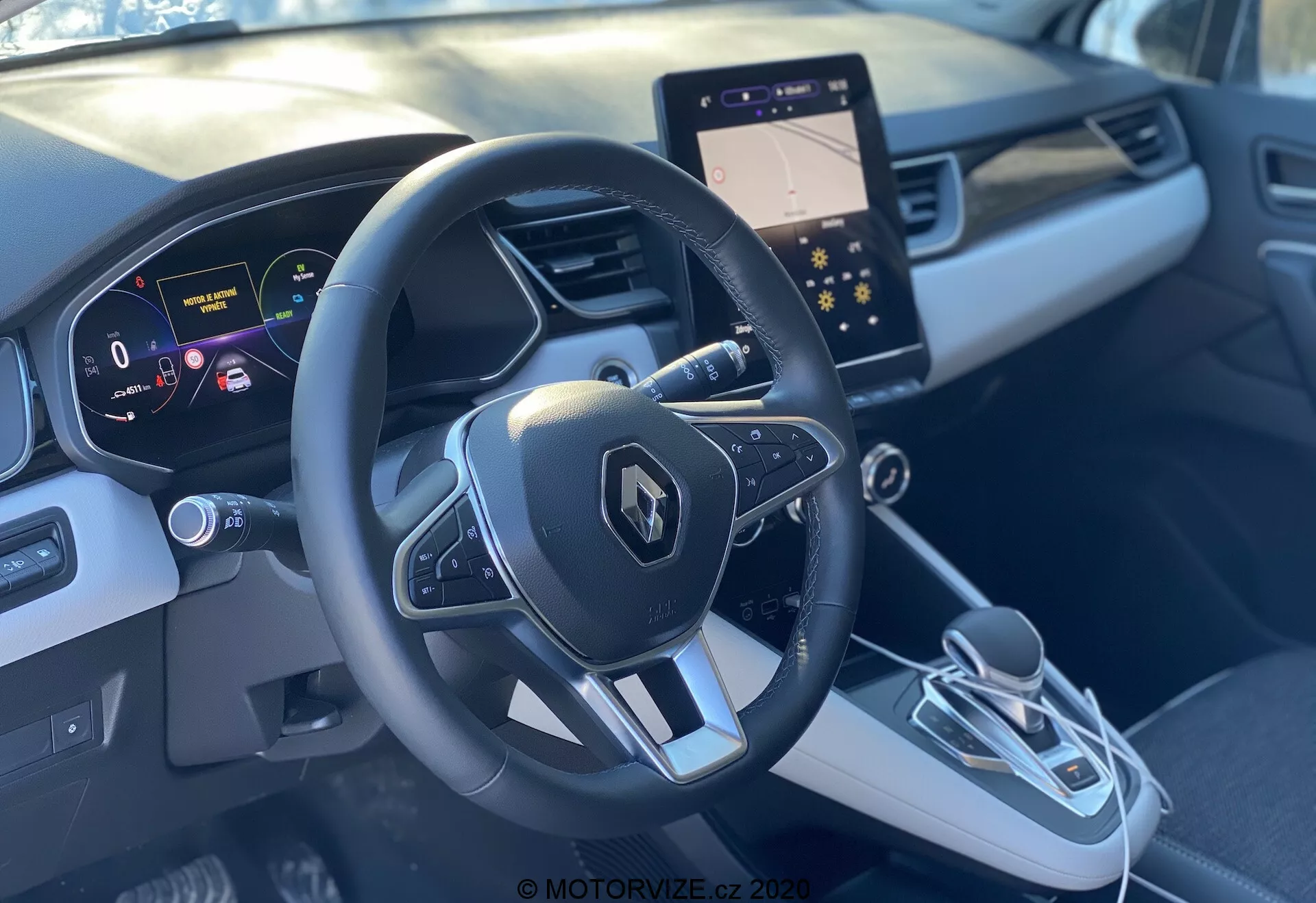 Pohľad do interiéru Renault Captur 2020 z miesta spolujazdca, ukazujúci športový trojramenný volant s logom Renault a multifunkčnými tlačidlami, digitálny prístrojový panel s čistým, vysokým rozlíšením zobrazujúci rýchlosť a navigačné pomôcky, a elegantnú stredovú konzolu. Stredová konzola obsahuje moderný infotainment displej, elektronickú radiacu páku a tlačidlo elektronickej parkovacej brzdy. Dvojfarebný šedý interiér s kontúrovaným dizajnom a ozdobnými lištami poskytujú prémiový pocit.