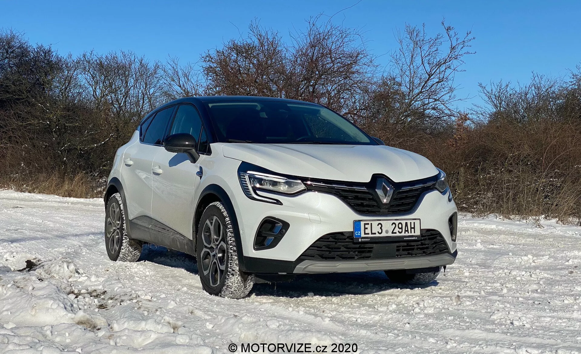 Biely Renault Captur 2020 zaparkovaný na zasneženom teréne pod jasnou oblohou, zobrazený z nízkeho predného trojštvrťového uhla s viditeľnou prednou mriežkou, svetlometmi s LED denným svietením, hmlovými svetlami, predným nárazníkom, zimnými pneumatikami, výrazným logom Renault, vyrezávanými linkami na kapote, zvýšenou svetlosťou, možnými senzormi parkovacej asistencie, bočnými zrkadlami a celkovým čistým, dobre udržiavaným stavom, s vodoznakom MOTORVIZE.cz © 2020.