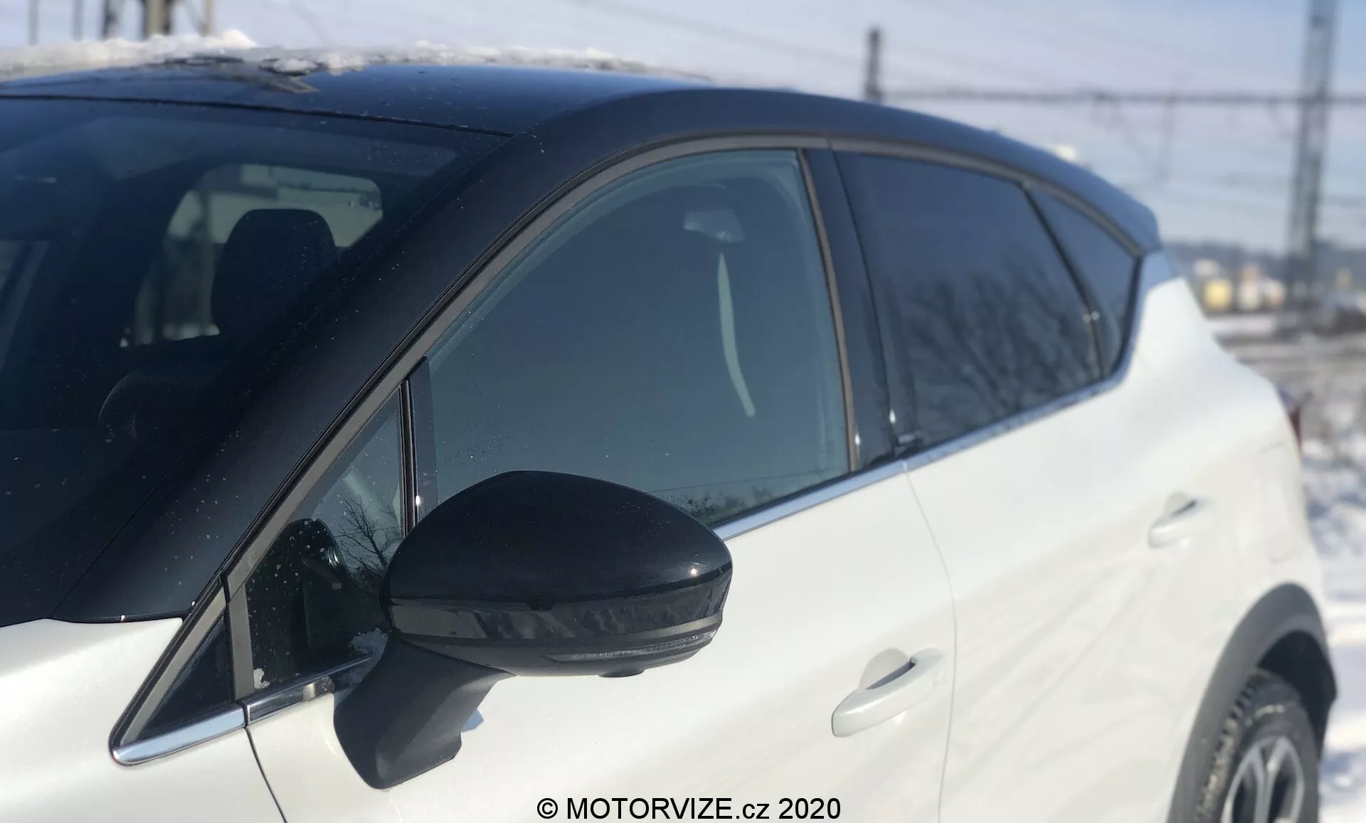 Zblízka bočný pohľad na biely Renault Captur 2020, zameraný od bočného zrkadla k priestoru pred predným kolesom. Výrazné čierne bočné spätné zrkadlo s integrovaným smerovým svetlom, fotené za denného svetla s prirodzeným slnečným svetlom. Mierne nízky uhol zdôrazňujúci strednú časť, plytká hĺbka ostrosti. Rozmazané pozadie s rozpoznateľnými prvkami ako plot, elektrické vedenie a štruktúru, naznačujúce vonkajšie prostredie. Lesklý biely lak vozidla a farebne zladené kľučky dverí, ukazujúce dobre udržiavaný moderný dizajn. Líniové okno stúpajúce k zadnej časti, ktoré naznačuje dynamický štýl.