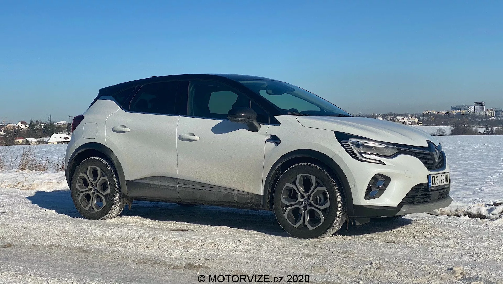 Renault Captur 2020 moderné crossover SUV umiestnené na ľadovom zasneženom teréne s jasnou oblohou v pozadí. Cestná strana smeruje ku kamere pod miernym uhlom, zobrazuje prednú, bočnú a čiastočne hornú časť vozidla. Obsahuje LED denné svietenie, integrovanú prednú mriežku s emblémom výrobcu, dvojfarebnú svetlú a čiernu farbu, päťramenné hliníkové kolesá, plastové ochranné lišty, elegantné zapustené kľučky dverí a zužujúcu sa líniu okien so zadným spoilerom. Naznačuje pokročilé technológie ako LED osvetlenie a bezkľúčový vstup, zasadené do chladného počasia blízko mestského prostredia.
