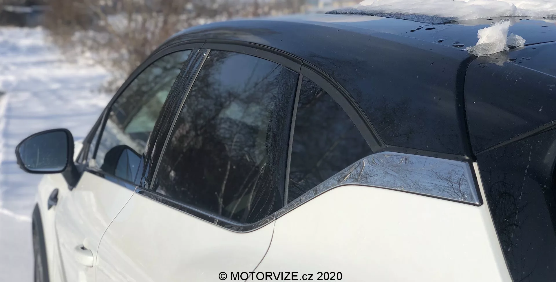Bočný pohľad na Renault Captur 2020 v zimnom prostredí, zvýrazňujúci čierno-bielu dvojfarebnú schému. Obrázok sa sústreďuje na B-stĺpik, časť zadných dverí a zadný panel, zachytený zo zadného uhlu. Snehové záplaty na streche a topiace sa ľad na okne odrážajú jasný deň.