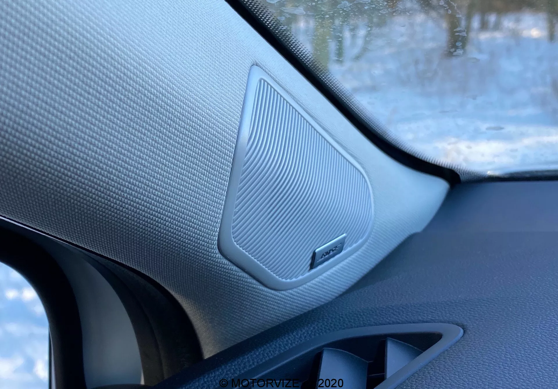 Detailný záber interiéru Renault Captur 2020, zobrazujúci reproduktor na predných dverách spolujazdca s textúrovanou mriežkou, obklopený kontrastnými materiálmi a emblémom značky, zhotovený vnútri vozidla.
