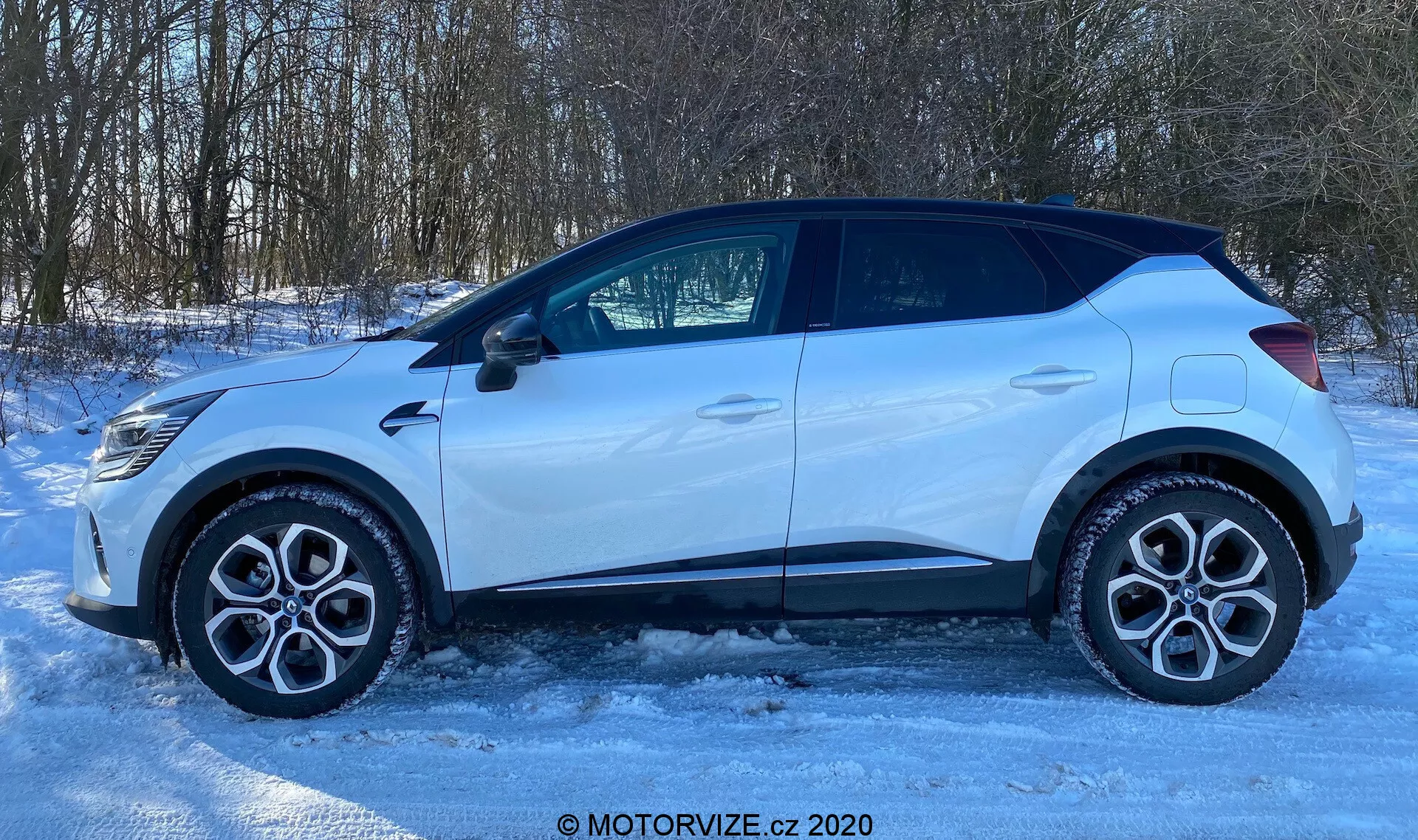 Bočný pohľad na Renault Captur 2020 zaparkovaný na zľadovatelej alebo zasneženej ploche. Profil vozidla ukazuje zliatinové kolesá, štandardné cestné pneumatiky, štyri dvere, bočné okná vrátane malého zadného okienka a bočné spätné zrkadlo s integrovaným indikátorom smeru. Strecha mierne klesá smerom k zadnej časti, spolu s plastovými obkladmi okolo blatníkov a spodnej časti dverí, čo naznačuje terénny dizajn. Dobre osvetlená scéna, pravdepodobne vďaka prirodzenému slnečnému svetlu, definuje tvar auta na pozadí vidieckeho alebo predmestského prostredia so stromami. Obrázok obsahuje vodoznak s nápisom 'MOTORVIZE.cz 2020'.