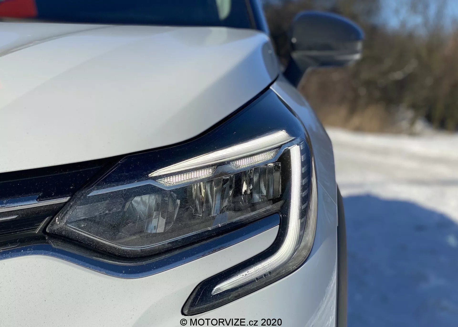 Detailný pohľad na predný ľavý roh moderného auta, zameraný na LED svetlomet a mriežku, vytvorený z nízkeho uhlu na úrovni svetlometu. Denné svetlá majú zakrivený dizajn objímajúci jednotku svetlometu. Viditeľné potenciálne adaptívne svetomety alebo rohové osvetlenie, s kovovým lakovaním a integrovanými smerovkami vo vonkajšom spätnom zrkadle. Jasné prirodzené osvetlenie naznačuje vonkajšie denné prostredie.