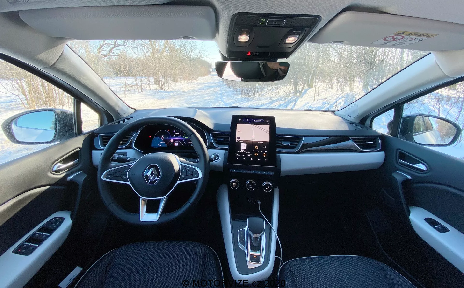 Pohľad na interiér Renaultu Captur 2020 zo zadného sedadla smerom dopredu, zobrazujúci jeho tmavé čalúnenie, centrálne spätné zrkadlo, osvetlený digitálny prístrojový panel za multifunkčným volantom, dotykový infotainment systém v stredovej konzole a ovládacie prvky pre elektrické okná, bočné zrkadlá, klimatizáciu a pravdepodobne strešné okno a tlačidlo SOS pre núdzové situácie.