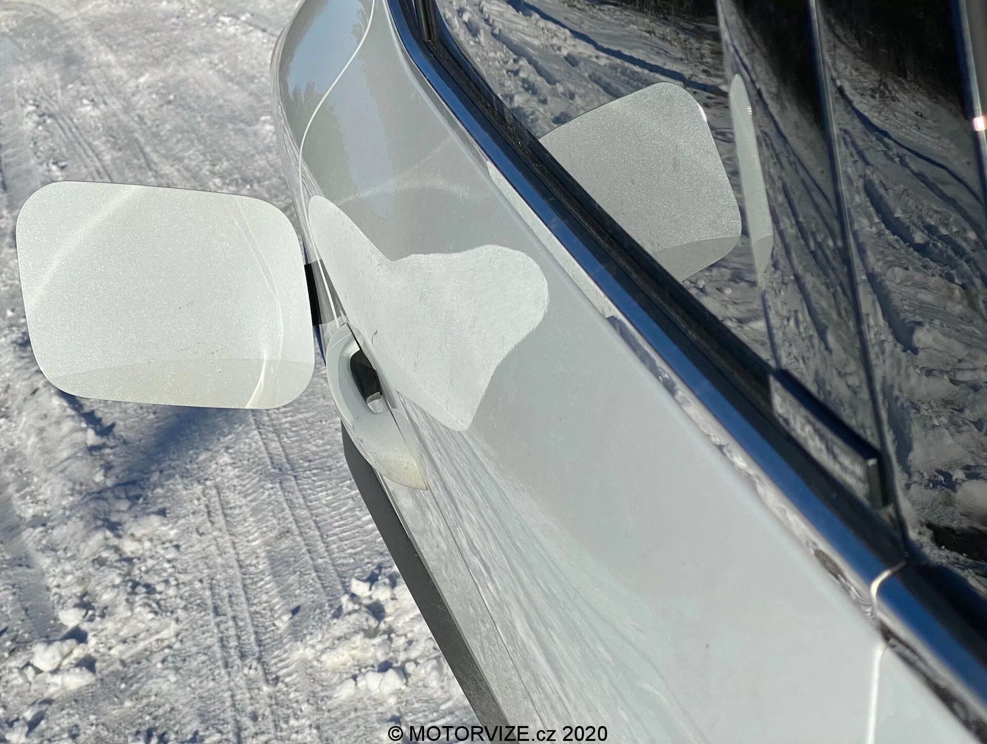 Detailný bočný pohľad na Renault Captur 2020 v zasneženom vonkajšom prostredí. Zameriava sa na pravé spätné zrkadlo, ktoré je zložené dopredu, čo môže naznačovať automatickú alebo manuálnu skladaciu funkciu. Na kryte zrkadla sú dve priehľadné ochranné fólie v tvare rovnobežníkov, pravdepodobne pre ochranu pri doprave alebo umývaní. Auto, ktoré sa zdá byť leskle tmavo čiernej alebo tmavo modrej farby, vyzerá dobre udržiavané s odrazmi naznačujúcimi čistý, leštený povrch. Okolitý terén a boky auta sú pokryté snehom. Viditeľná je aj časť kľučky predných dverí s tlačidlom alebo snímačom bezkľúčového prístupu.