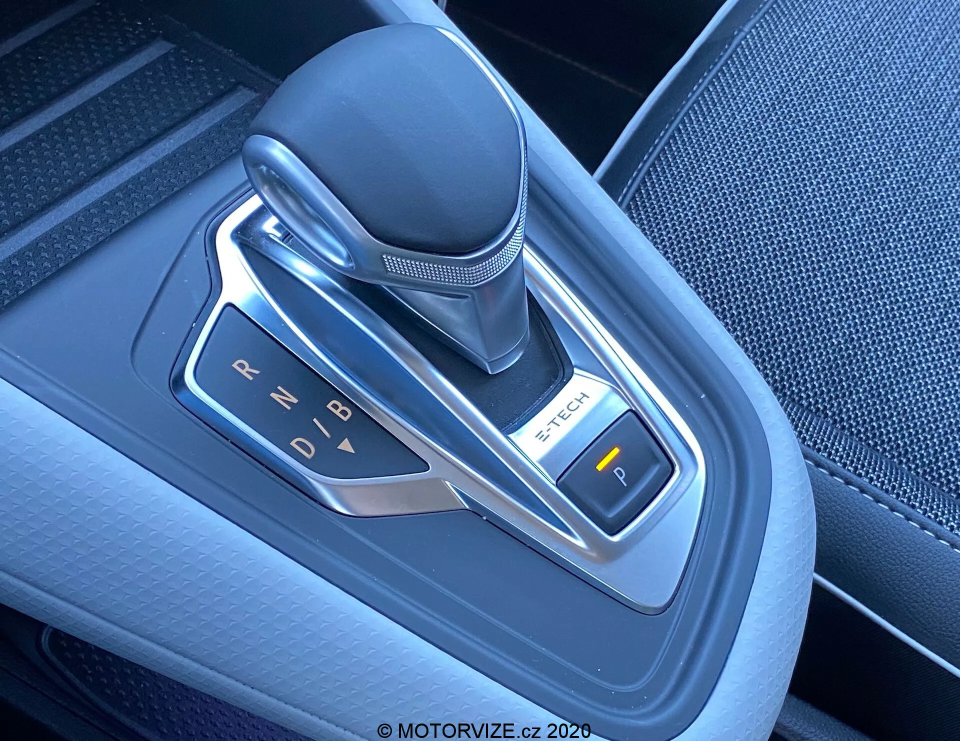 Detailný záber na modernú automatickú radiacu páku v Renault Captur 2020, umiestnenú na stredovej konzole. Elektronický výber rýchlostí je vertikálne orientovaný a vyrobený z kože, plastu a kovových akcentov. Fotografia, urobená zo zhora mierne z pravého uhla, zdôrazňuje označenia rýchlostí (P, R, N, D, B) osvetlené LED diódami. Na pravej strane je viditeľné tlačidlo s označením 'P', ktoré pravdepodobne slúži ako elektronická parkovacia brzda. Oblasť okolo radiacej páky je vyrobená z kvalitného textúrovaného plastu a kovových akcentov, čo odráža pokročilé technológie a moderný dizajn interiéru vozidla.