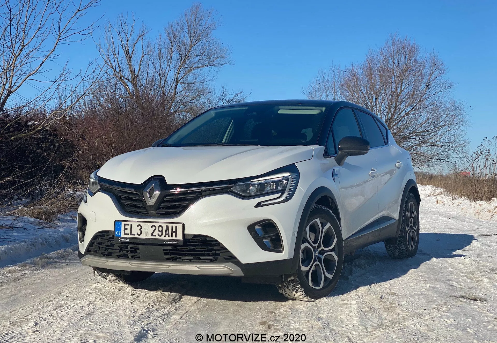 Moderné hatchback vozidlo Renault Captur 2020 zachytené z nízkeho predného trojštvrtového uhla, ktoré ukazuje prednú mriežku, kapotu, LED svetlomety, predný nárazník s integrovanými hmlovými svetlami a čiastočný bočný profil. Dizajnové prvky zahŕňajú výraznú mriežku s emblémom výrobcu, tvarované línie kapoty a viacramenné zliatinové kolesá, to všetko pod jasnou dennou oblohou, odrážajúc športový a aerodynamický vzhľad, možno vyznačujúci vylepšenú výbavu alebo športový balík. Auto je umiestnené na zasneženej ceste, čo naznačuje vlastnosti vhodné na zimnú jazdu.