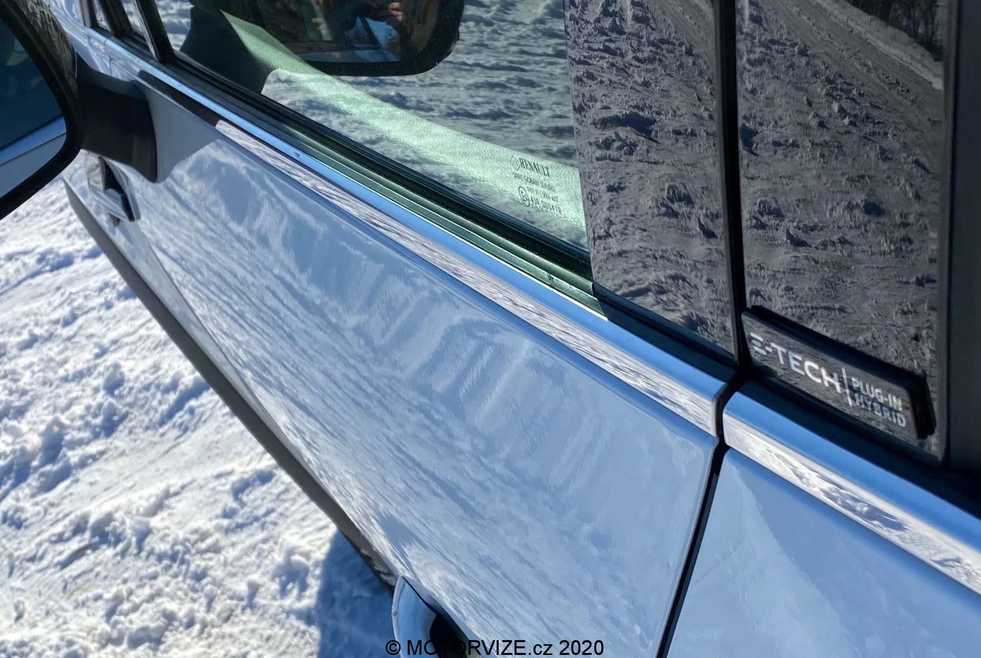 Detailný záber na Renault Captur 2020 v metalickej modrej farbe zameraný na oblasť dverí vodiča a bočného zrkadla s integrovaným smerovkou. Obrázok zvýrazňuje oblasť dverí s čiernym okrajom pozdĺž okraja tónovaného okna, ktorý odráža okolité prostredie. Na dverách je čiastočne viditeľný nápis označujúci možné vybavenie alebo technologický balík.