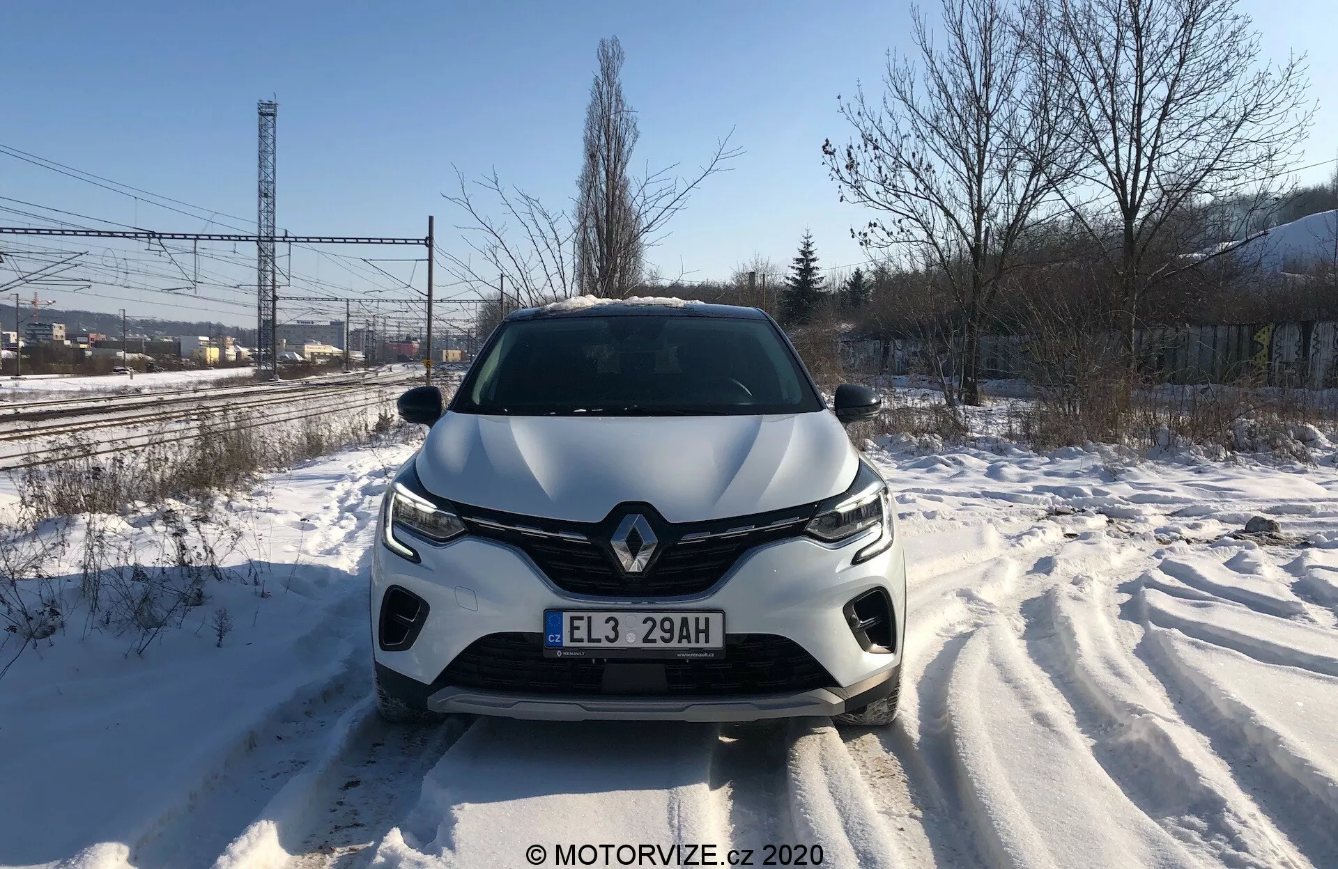 Priamy predný pohľad na Renault Captur 2020 na zasneženej ceste, ktorý počas dňa detailne zobrazuje prednú mriežku, svetlomety, tvarovanú kapotu a predný nárazník. Viditeľné stopy pneumatík svedčia o nedávnom pohybe. Dizajn vozidla naznačuje aerodynamické tvarovanie, s výrazným emblémom značky a pokročilým osvetľovacím systémom. Do predného nárazníka môžu byť integrované senzory alebo kamery asistencie pre vodiča.