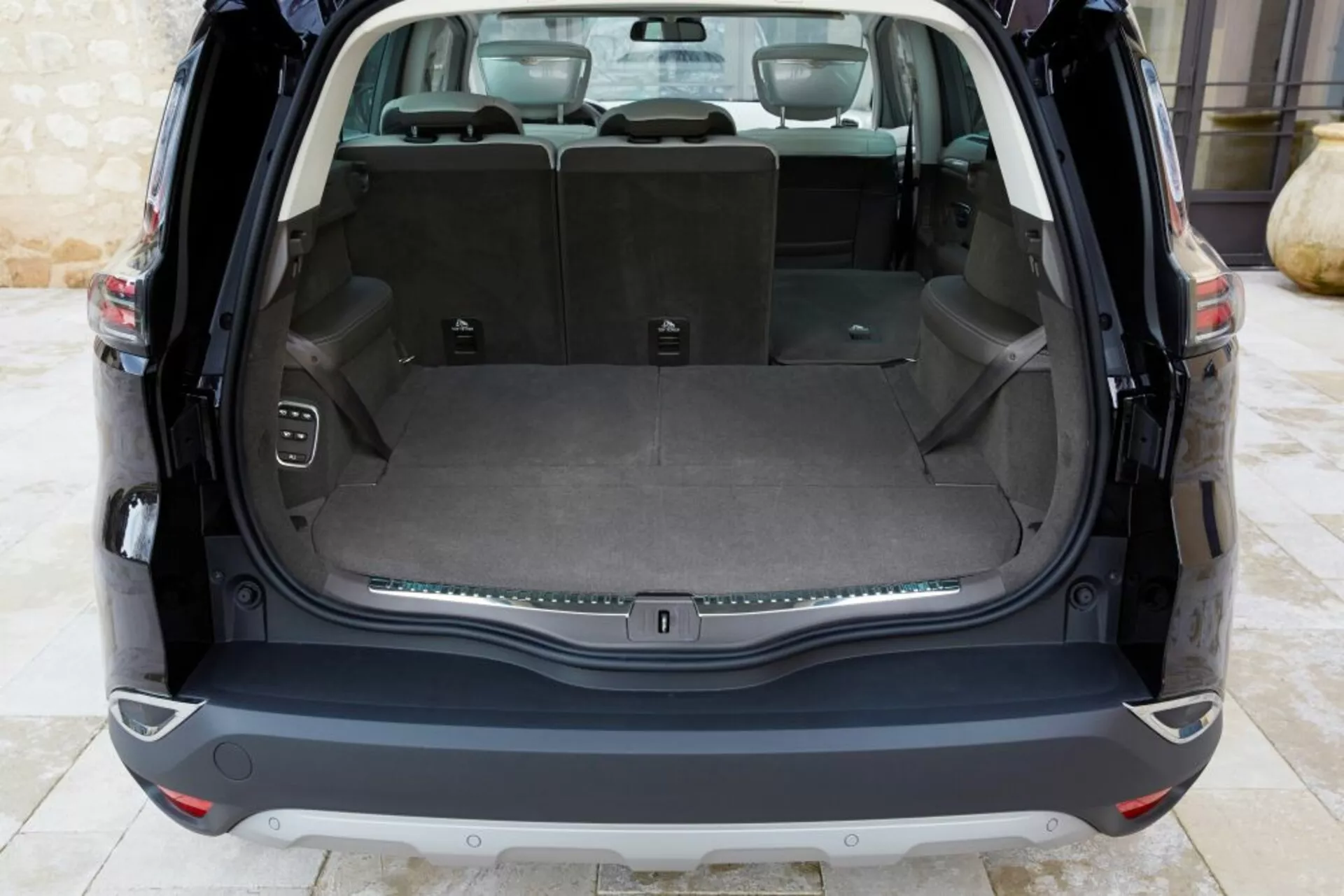 Zadný interiérový pohľad na Renault Espace 2015 s čistou nákladnou oblasťou a otvoreným vekom, ktorý obsahuje plochú nákladnú plochu, zadné sedadlá delené v pomere 60/40, elektrickú zásuvku, úchyty na náklad, tlačidlo ovládania elektronického zadného veka a chrómovú ozdobnú lištu, v prírodnom osvetlení.