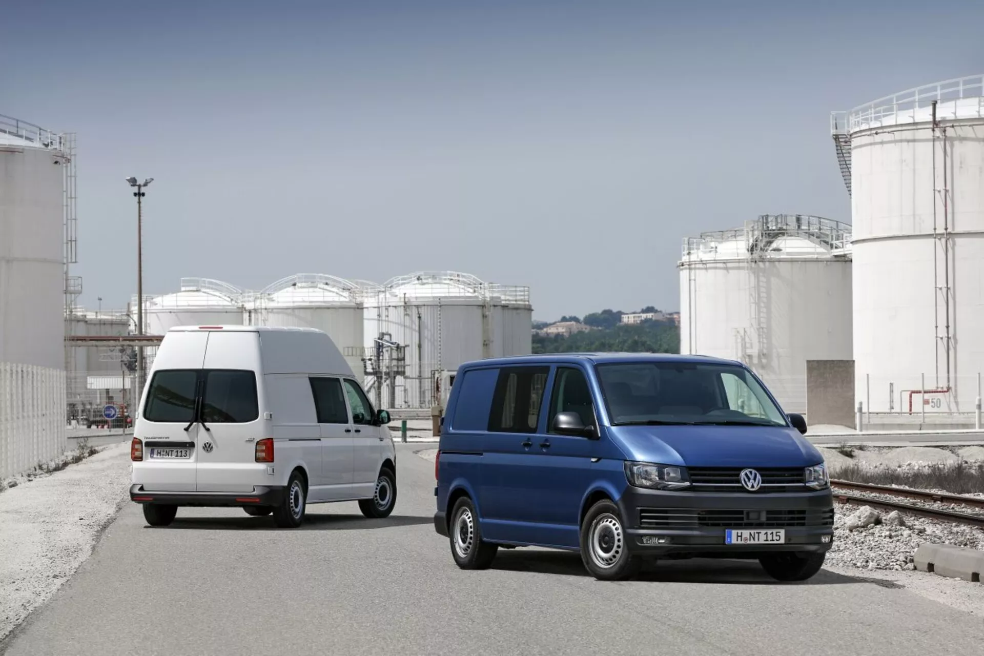 Dve dodávky Volkswagen Transporter zaparkované na priemyselnom mieste s veľkými úložnými nádržami v pozadí, ukazujúce ich komerčný úžitkový dizajn, posuvné bočné dvere a štandardné zliatinové kolesá.
