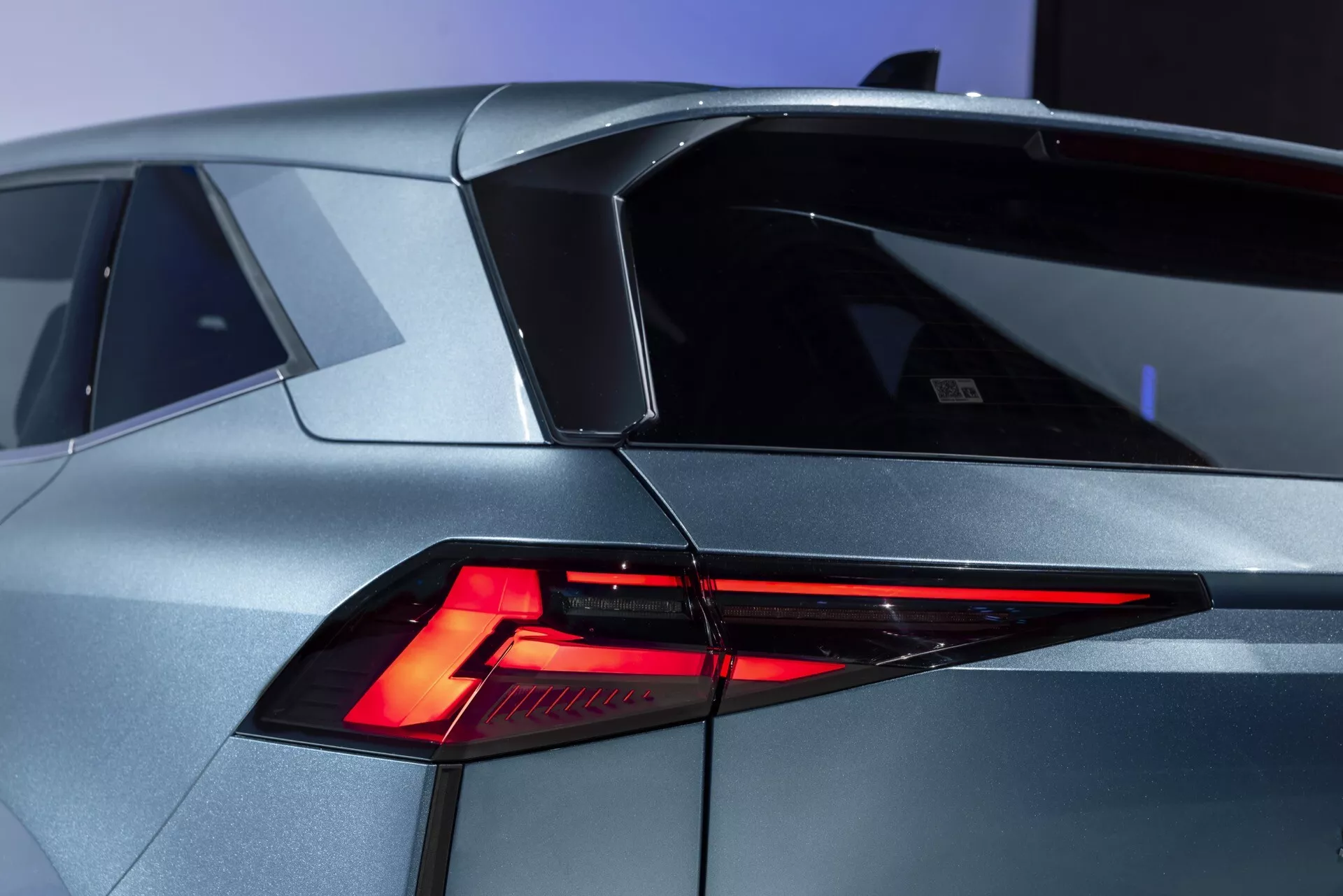 Zblízka moderného Renault Symbioz 2024 (1. generácia) z trojštvrťového zadného uhla. Obraz výrazne zobrazuje ostrý, uhlový dizajn LED zadných svetiel, sklonené zadné okno a charakteristickú líniu strechy s malým spojlerom. Karoséria ukazuje futuristické geometrické tvary v kovovom striebornom/sivom prevedení. Na ľavej strane kufra je viditeľný znak alebo emblém, naznačujúci model alebo značku auta. Ďalšie detaily zahŕňajú časť, ktorá pripomína uzáver palivovej nádrže na ľavom zadnom paneli.